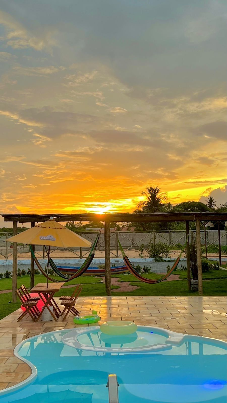 Uma piscina com redes, mesas, cadeiras e guarda-sóis ao pôr do sol.