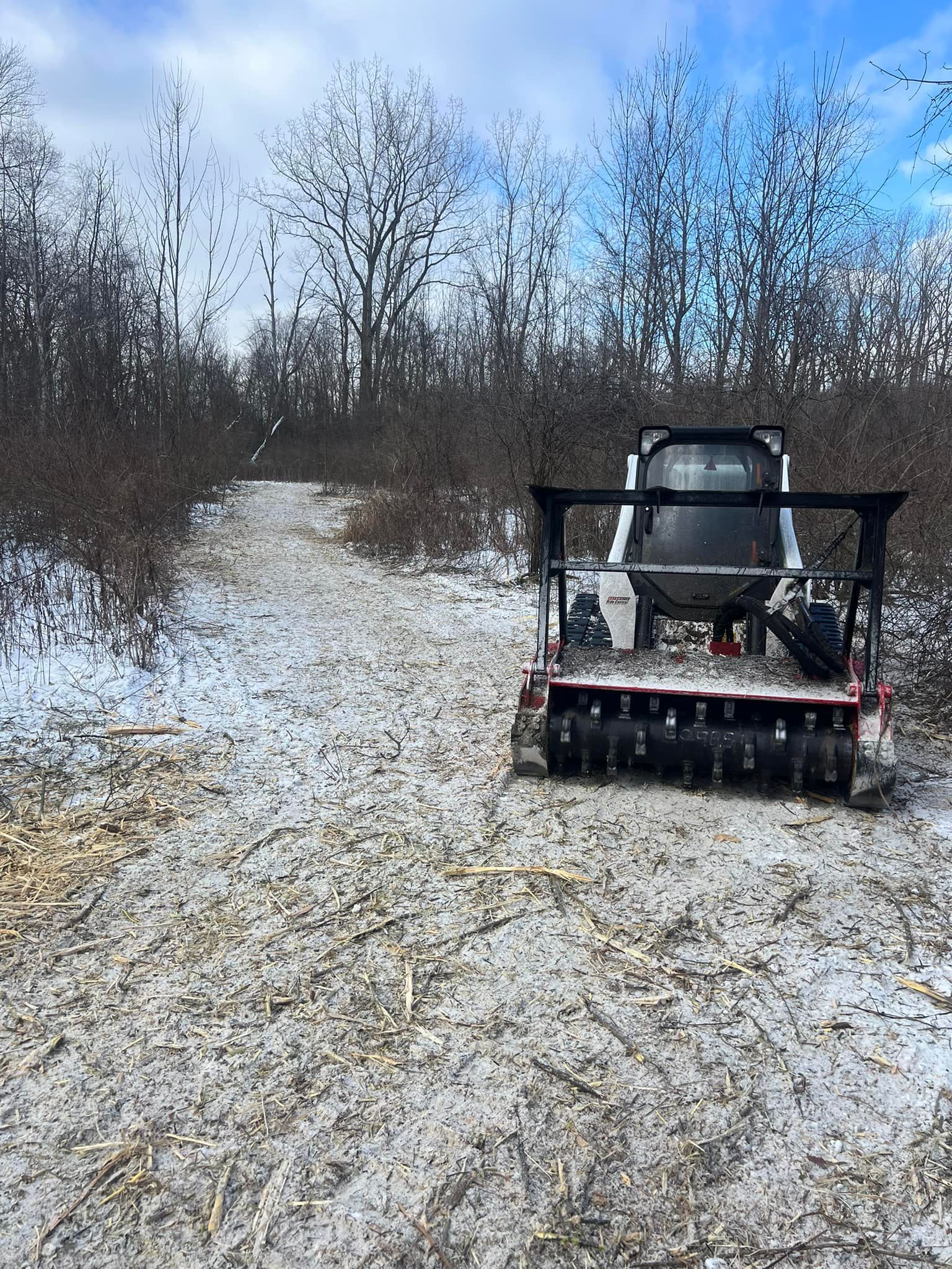 Skid steer mulcher clearing a snowy trail through a forest.