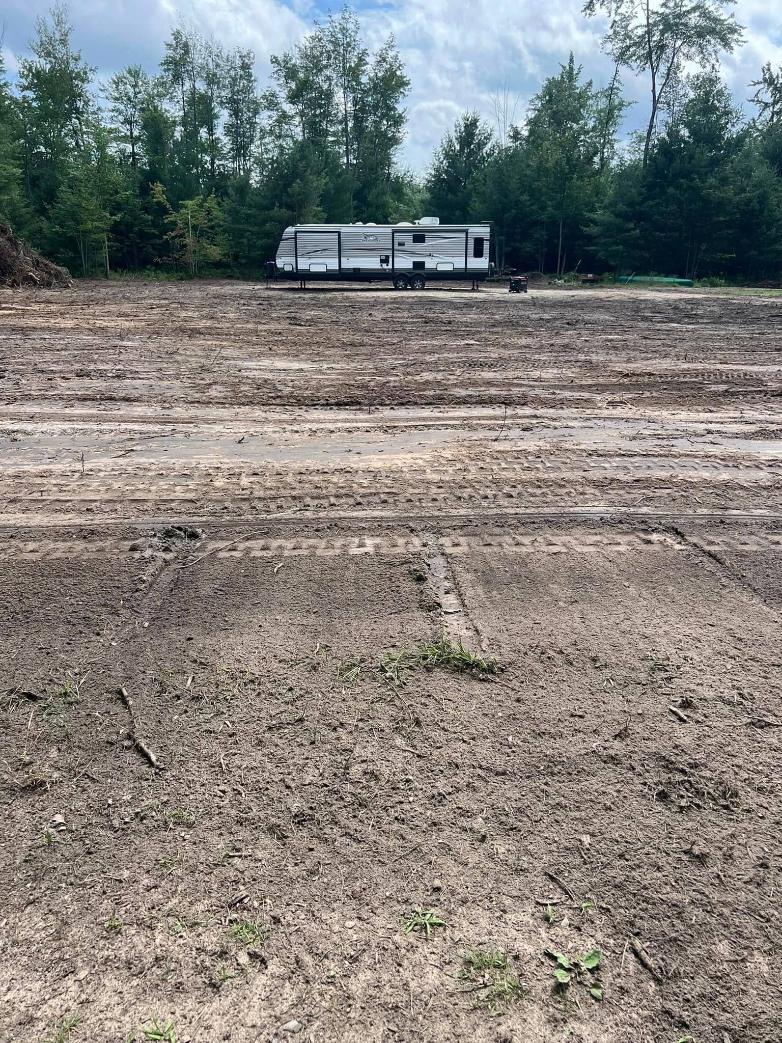 A travel trailer parked on a muddy, cleared lot, with trees in the background under a cloudy sky.