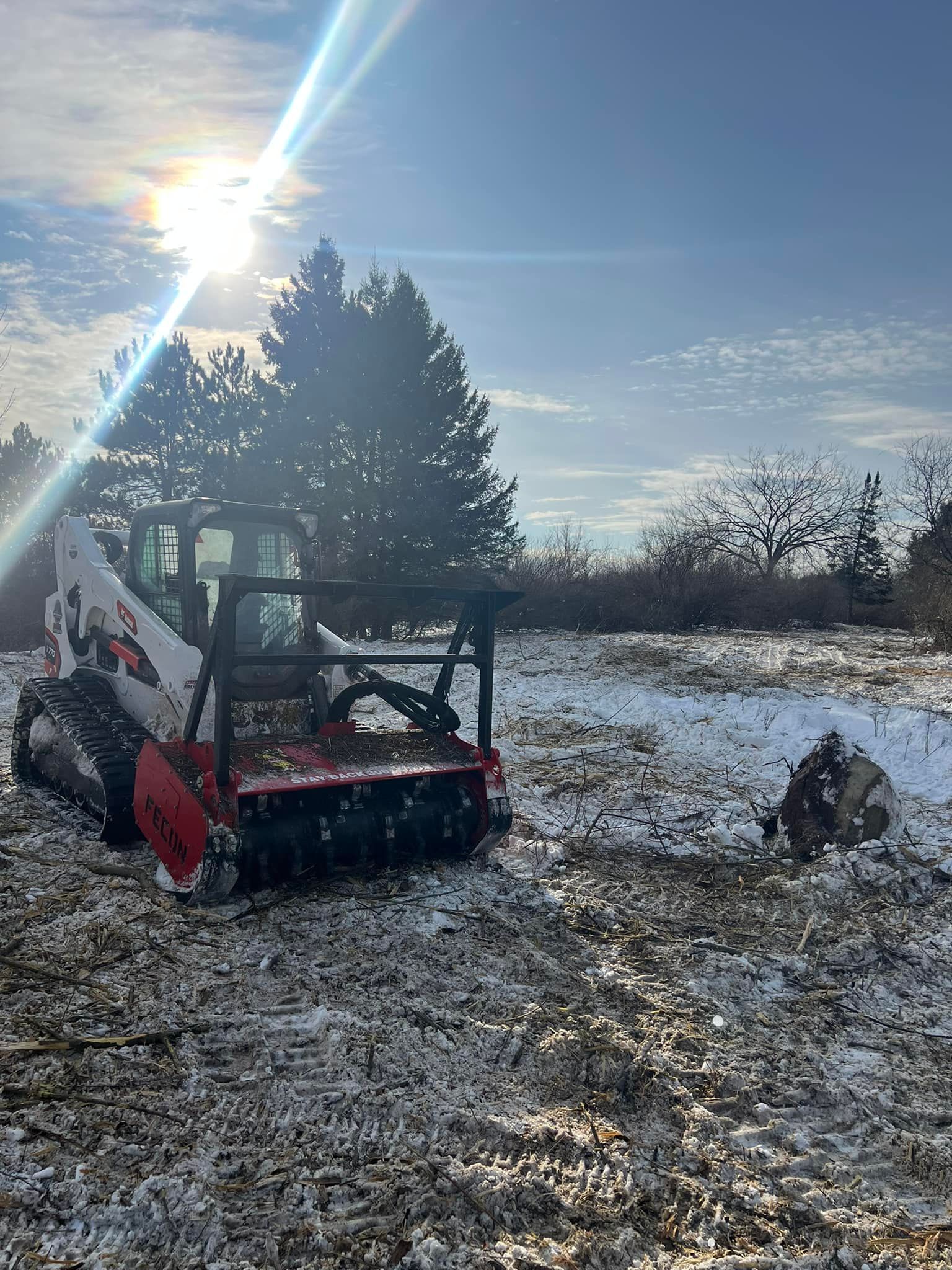 Bobcat with grinder attachment clearing land, partial snow cover, bright sun.