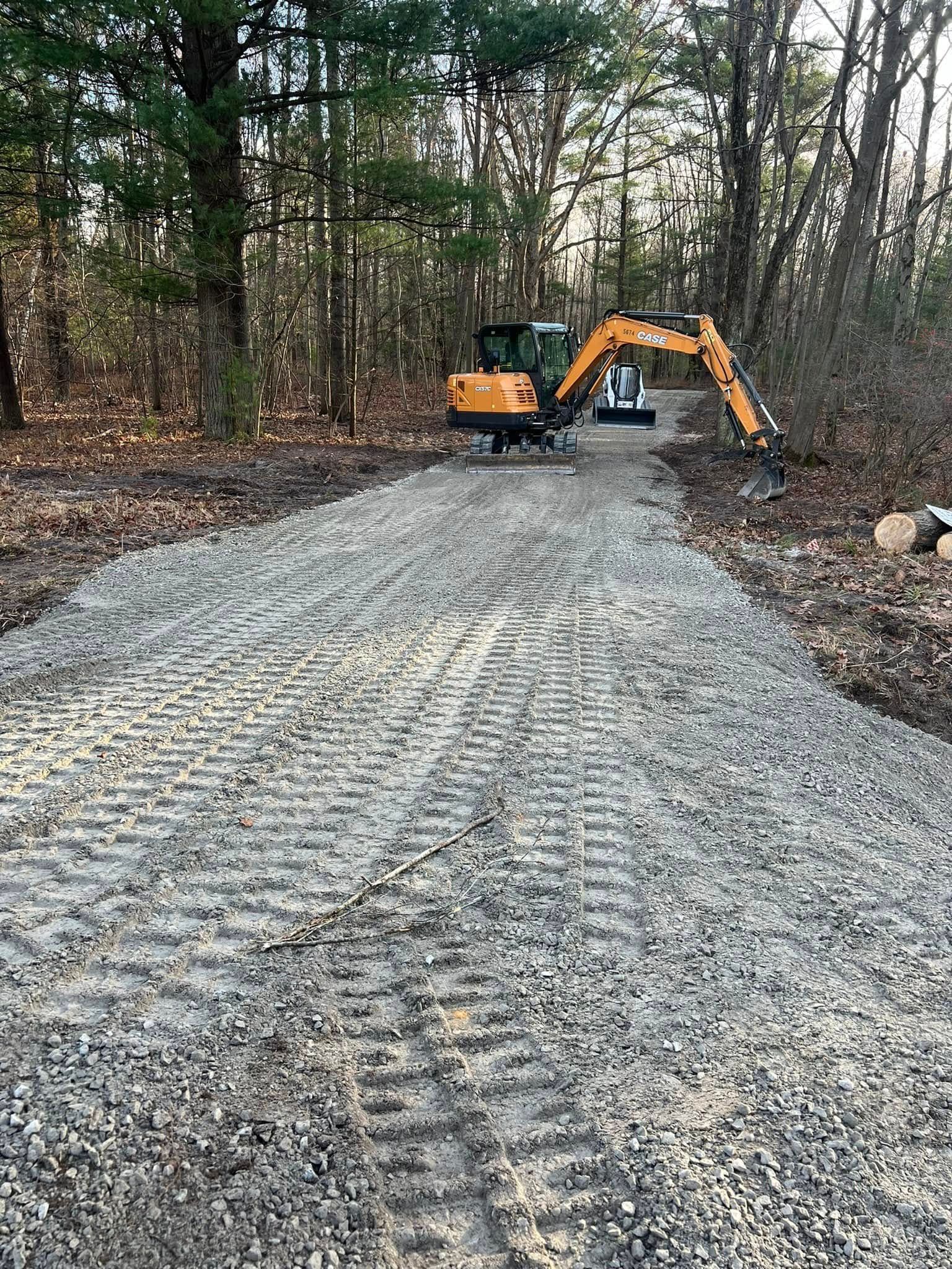 Excavator on a gravel road in a wooded area, grading the surface.