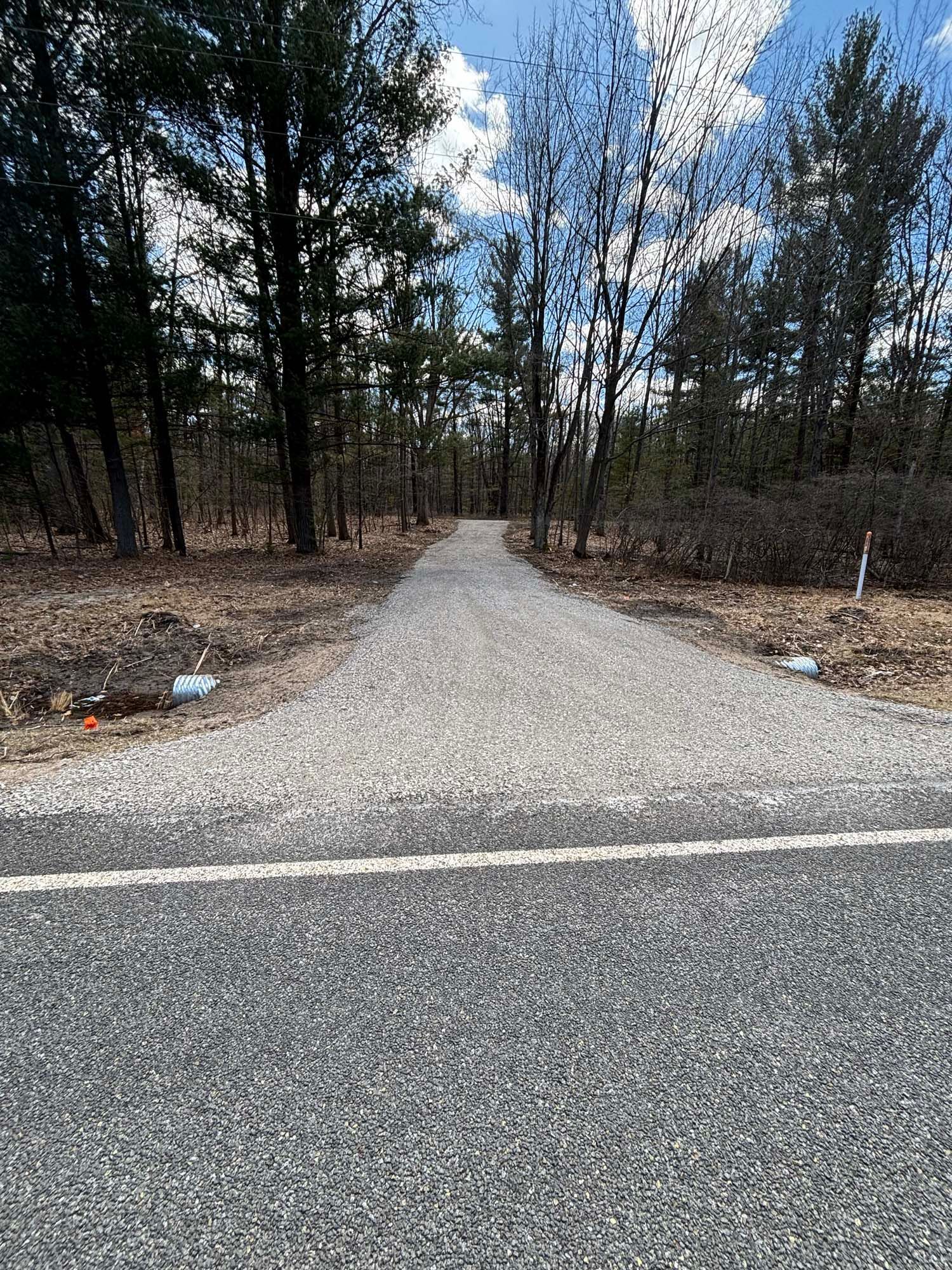 Gravel path leading into a forest, emerging from a paved road. Trees line the path on either side under a cloudy sky.