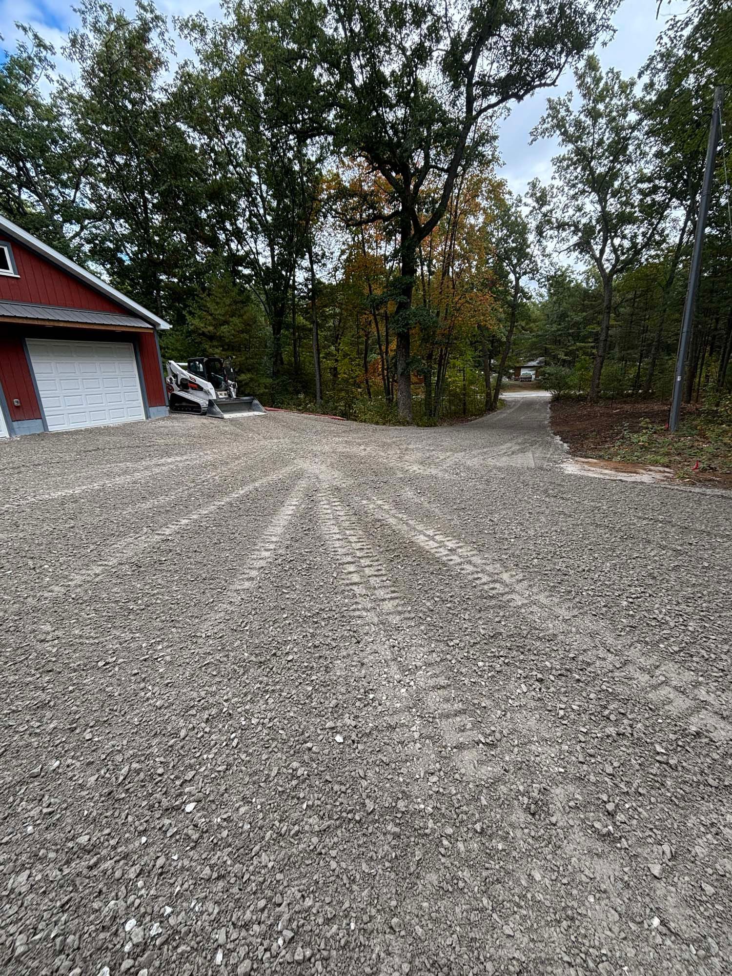 Gravel driveway forks, leading to a red building with a white garage door and a wooded area. Cloudy sky.
