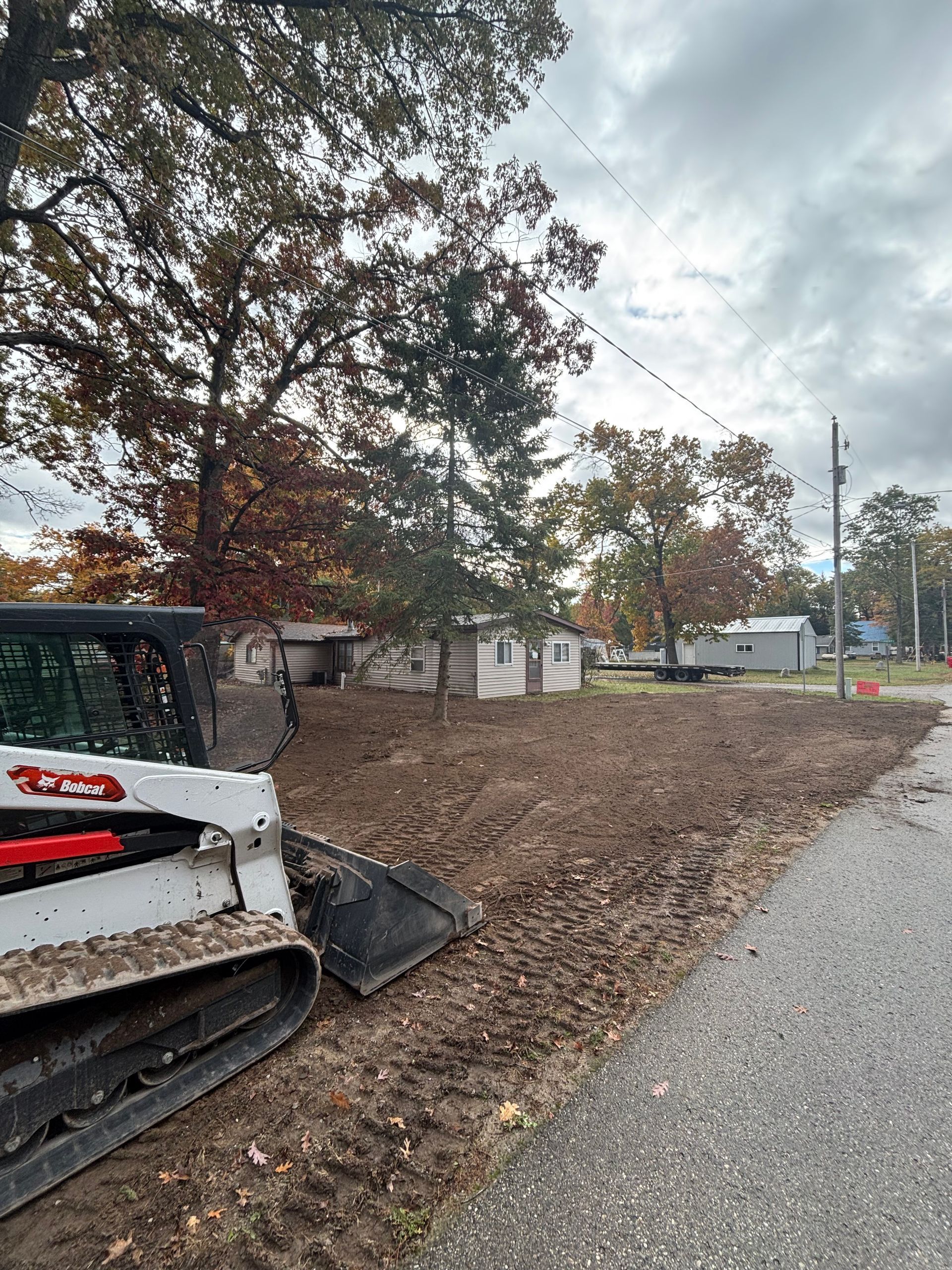 Bobcat skid steer next to a patch of dirt and fallen leaves, roadside construction in front of trees.