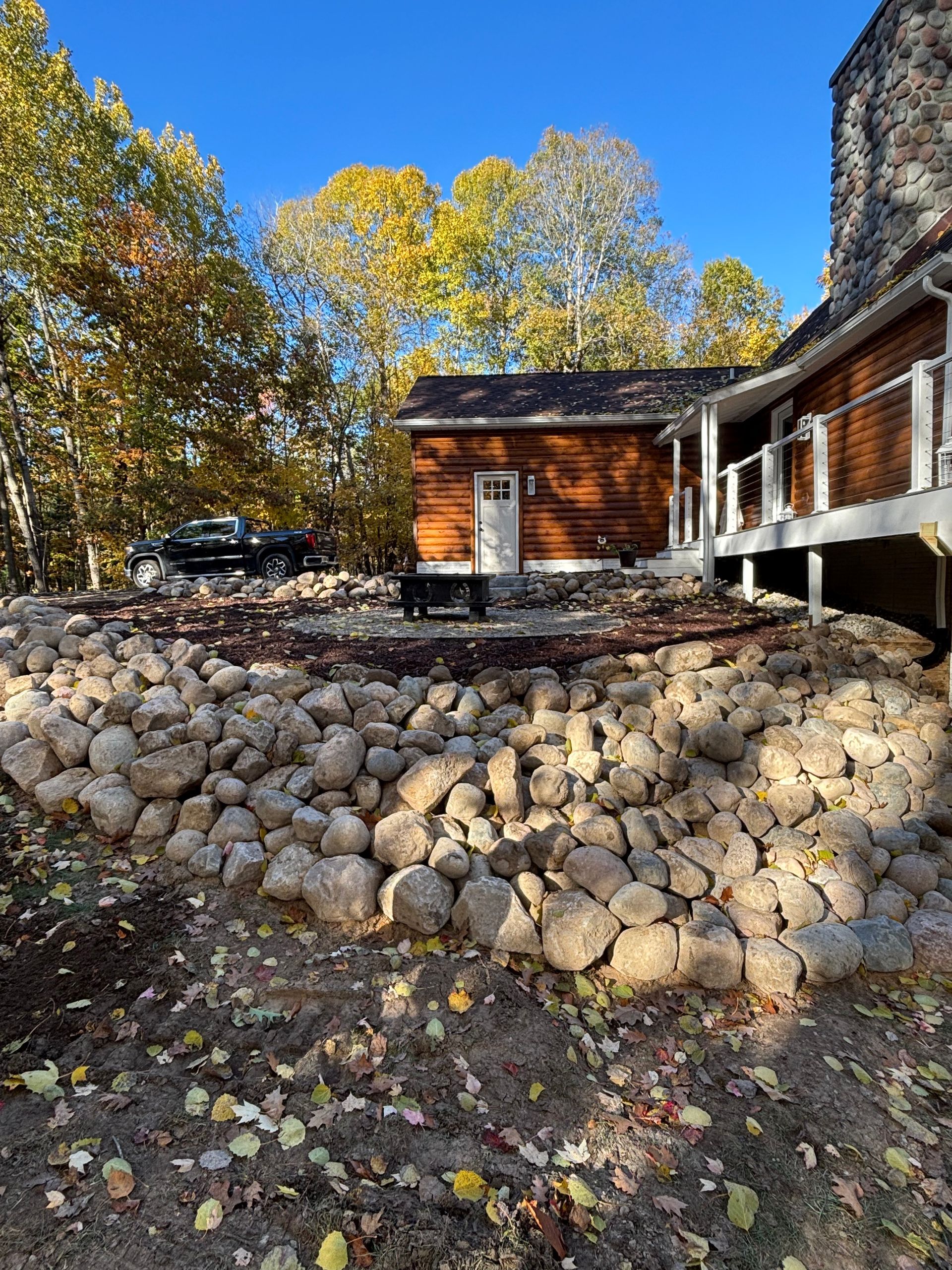 Rocks and dirt form a retaining wall in front of a wood building with a porch under a blue sky, trees in the background.