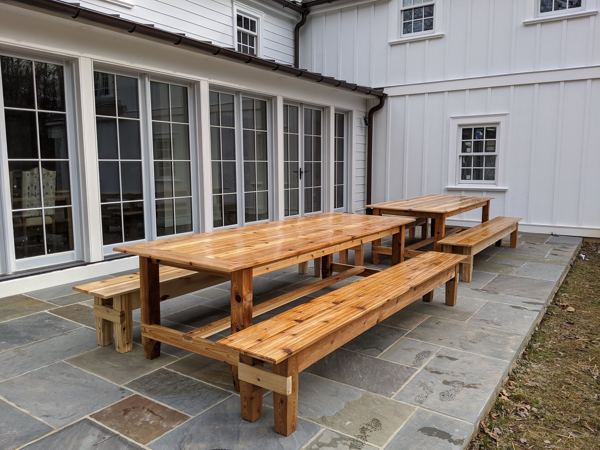 A large wooden picnic table and benches on a patio in front of a white house.