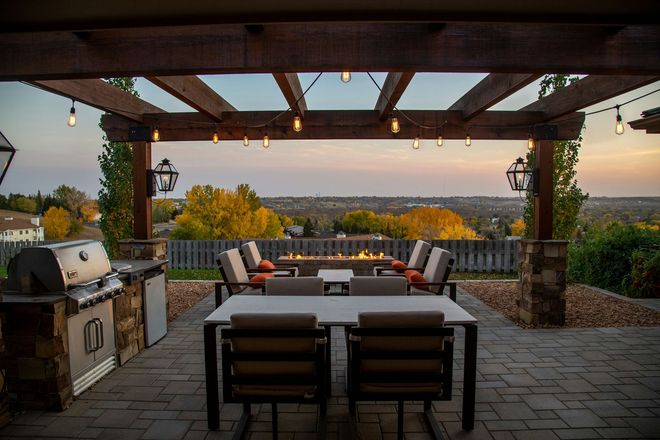 Outdoor patio with dining table, grill, and fire pit, under a pergola with string lights, overlooking a cityscape at sunset.