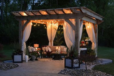 A cozy outdoor pergola with string lights, white curtains, and a seating area illuminated at dusk.