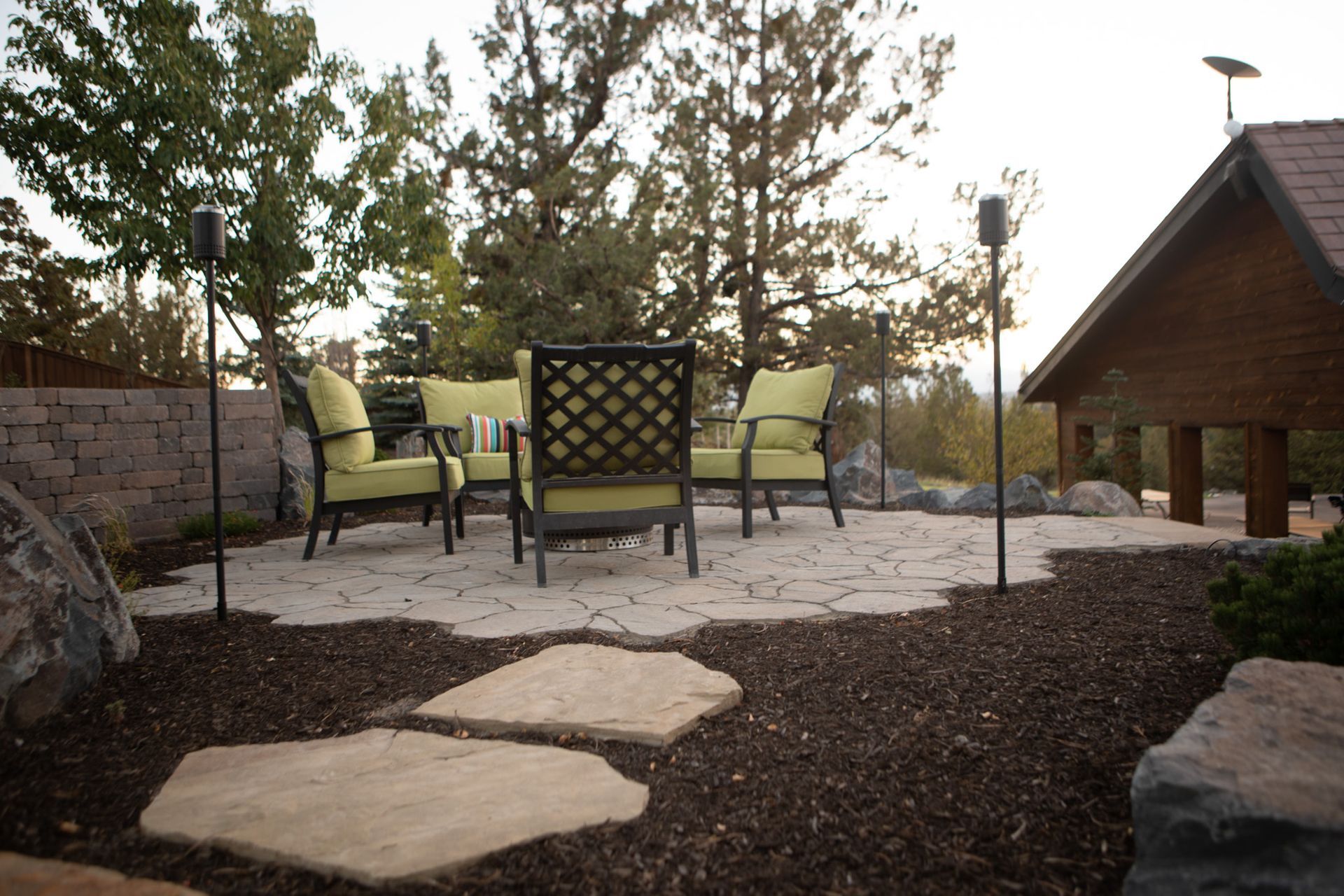Outdoor patio with green cushioned chairs, stone pavers, and a wooden structure, at dusk.