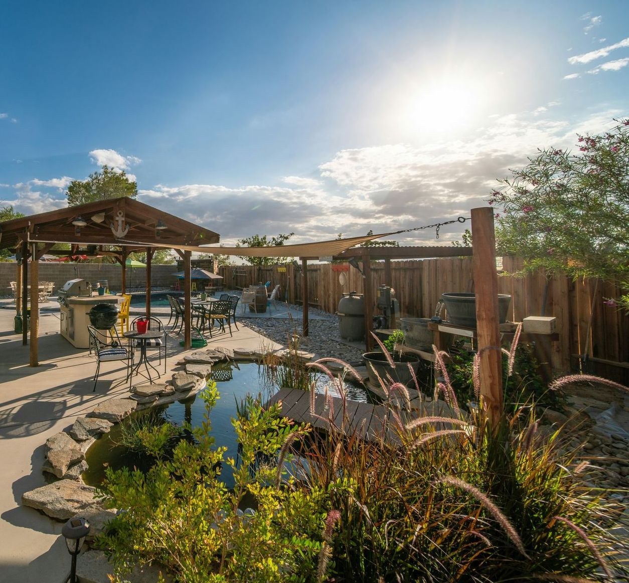 Backyard with pond and seating under a pergola, surrounded by plants and a wooden fence, sunny day.