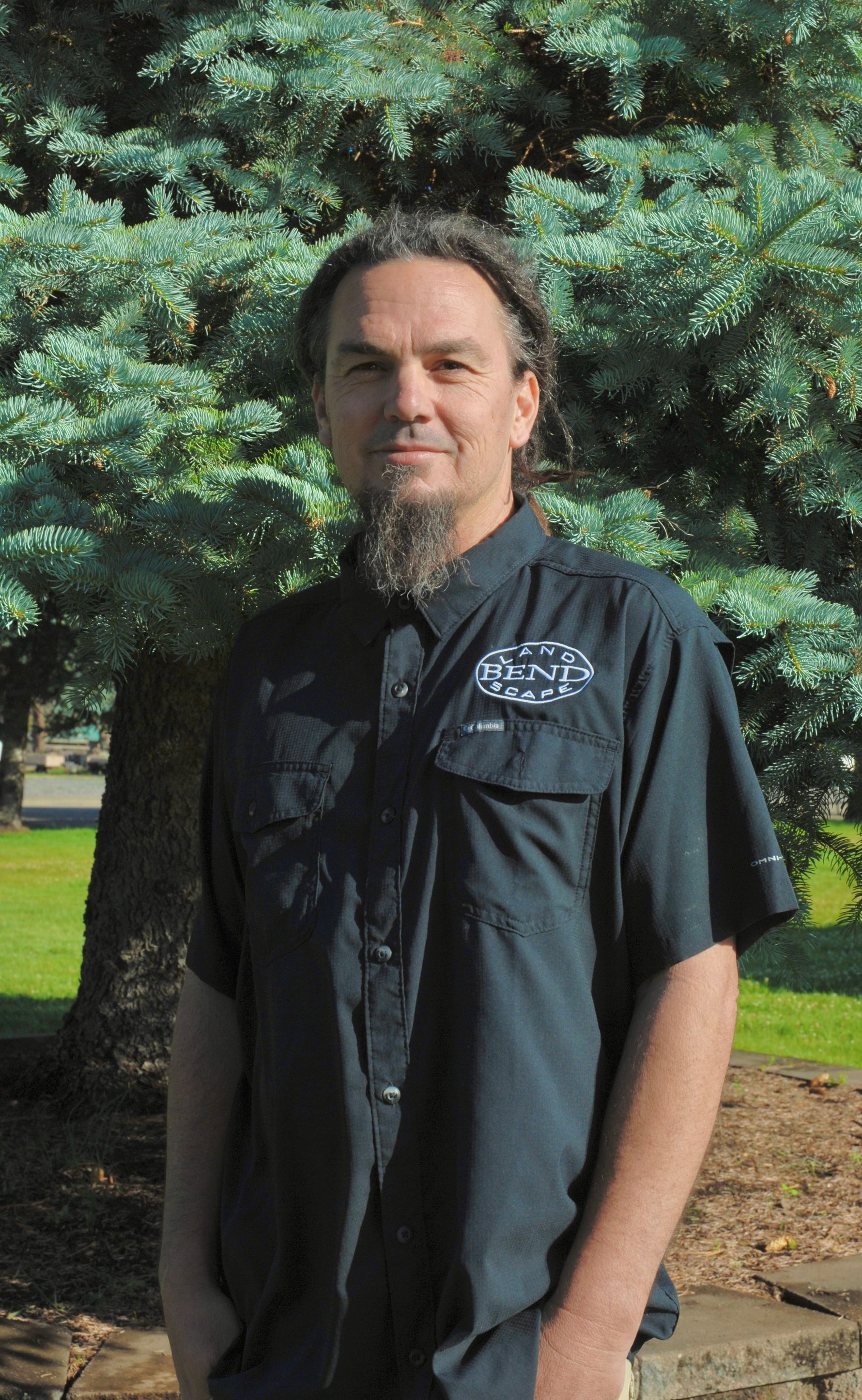 Man with a beard and long hair in a black shirt stands in front of a green bush on a sunny day.