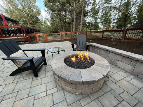 Two black Adirondack chairs face a lit fire pit on a brick patio, with a glass-topped table. A brick wall and fence create the backyard setting.