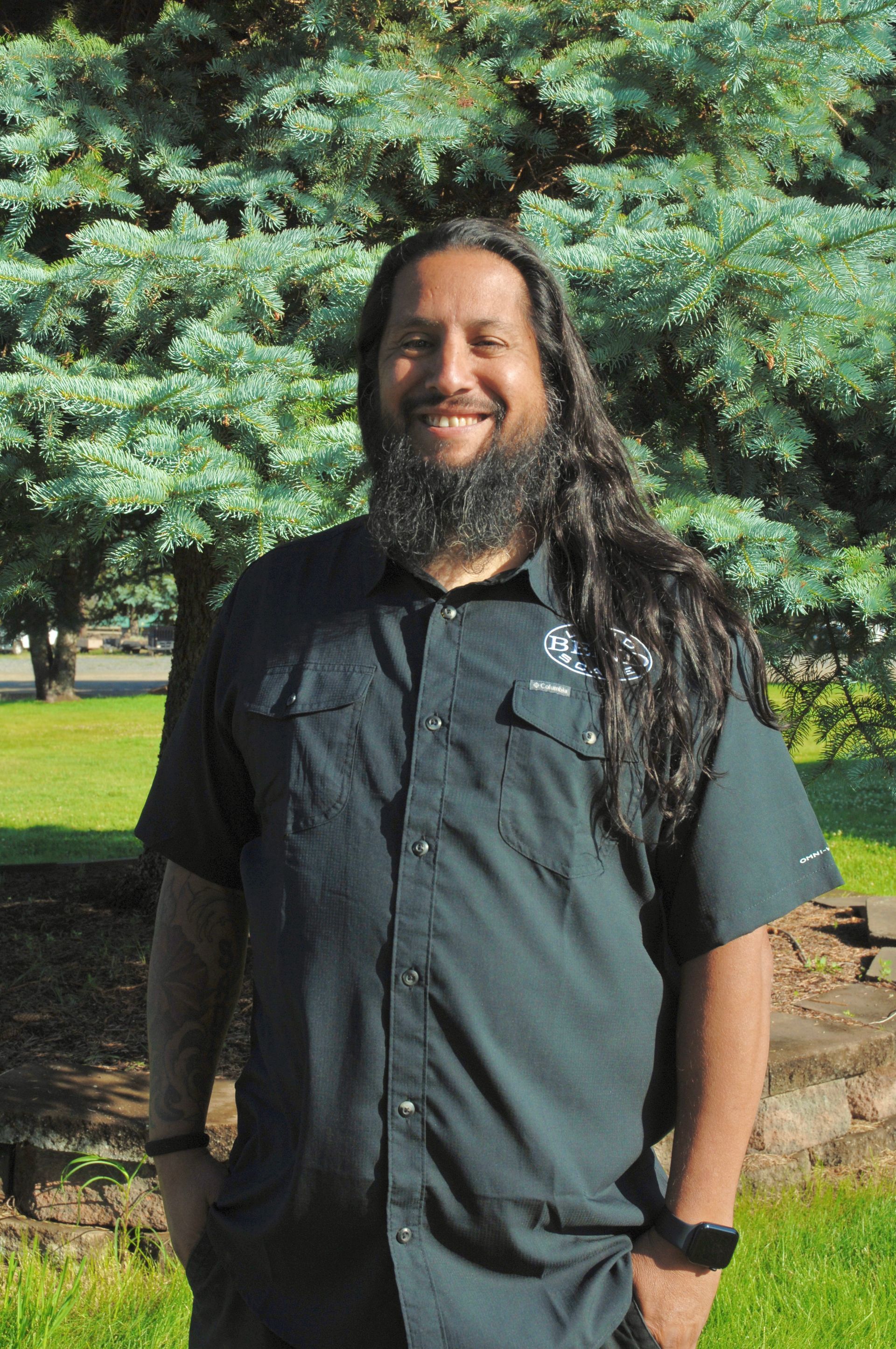 A smiling man with long, dark hair and a beard stands outside, wearing a dark button-down shirt.
