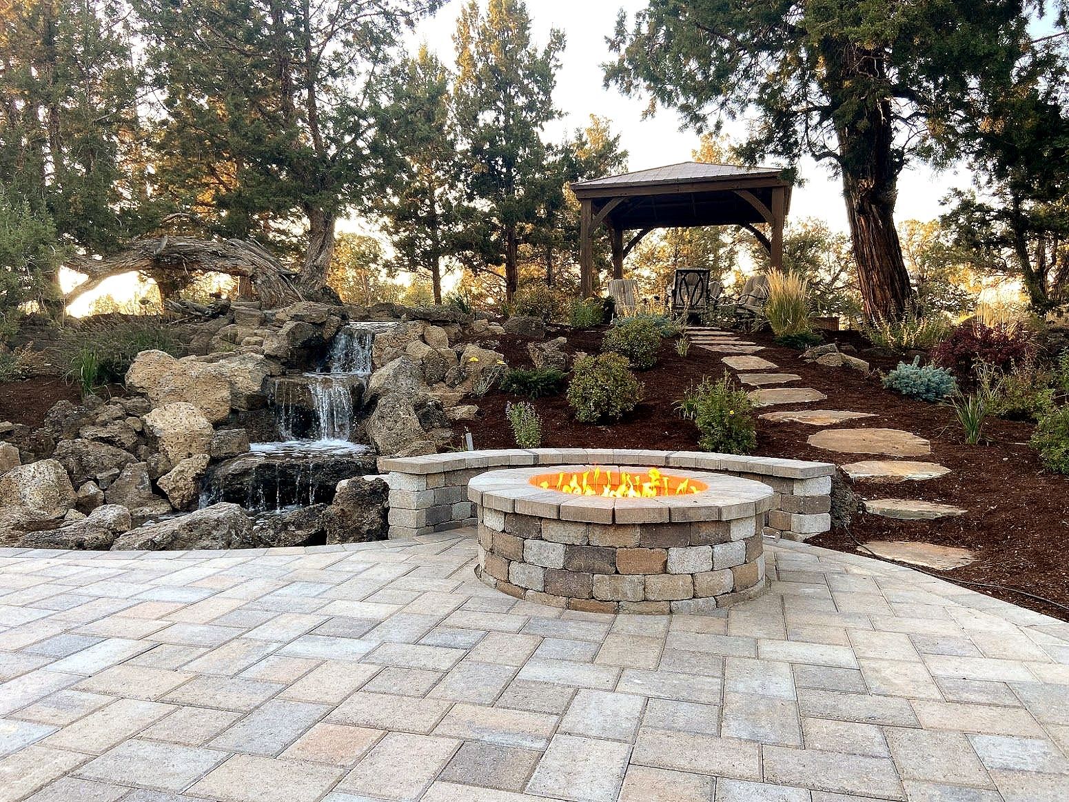 A backyard with a brick patio, a fire pit, and a small waterfall. A gazebo sits atop stone steps.