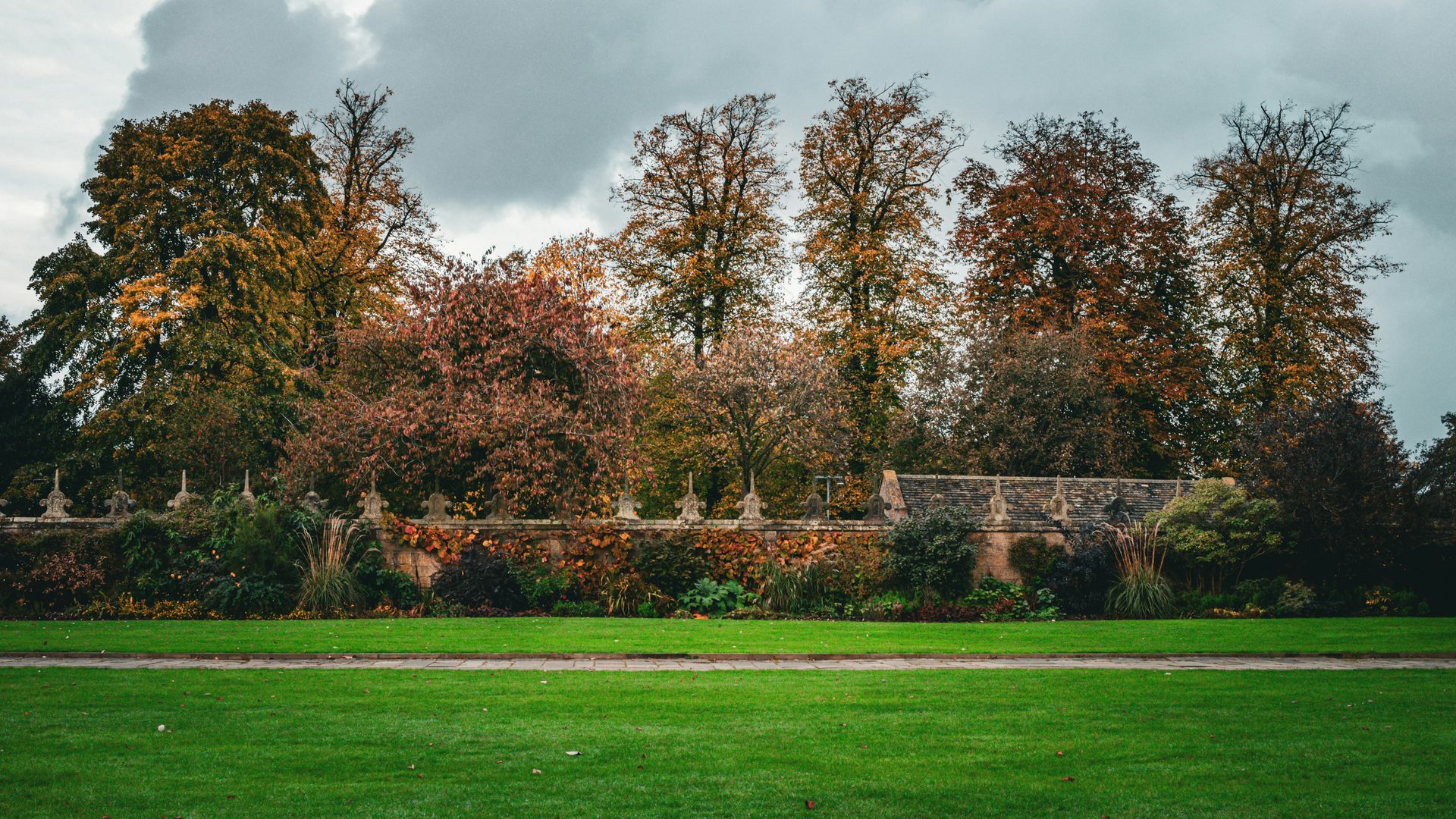 Green lawn in foreground, a brick wall with shrubs and colorful fall trees against a cloudy sky.