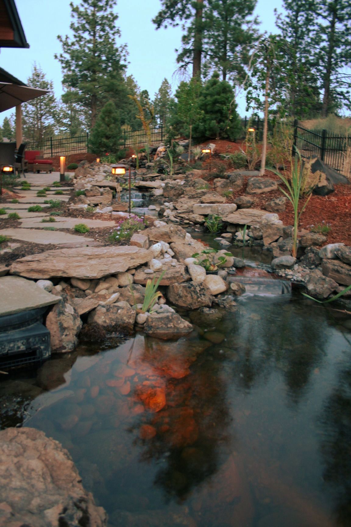 A landscaped pond with a waterfall feature. Rocks and greenery surround the water, reflecting the evening sky.