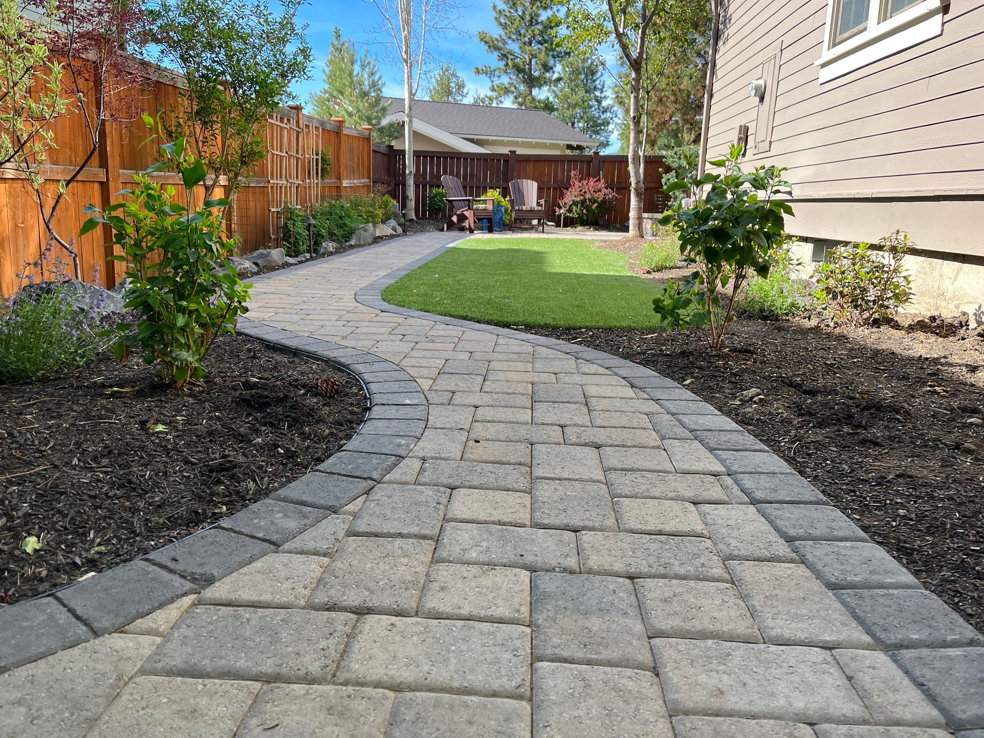 Brick pathway curves through a backyard with a small lawn, trees, and a wooden fence.