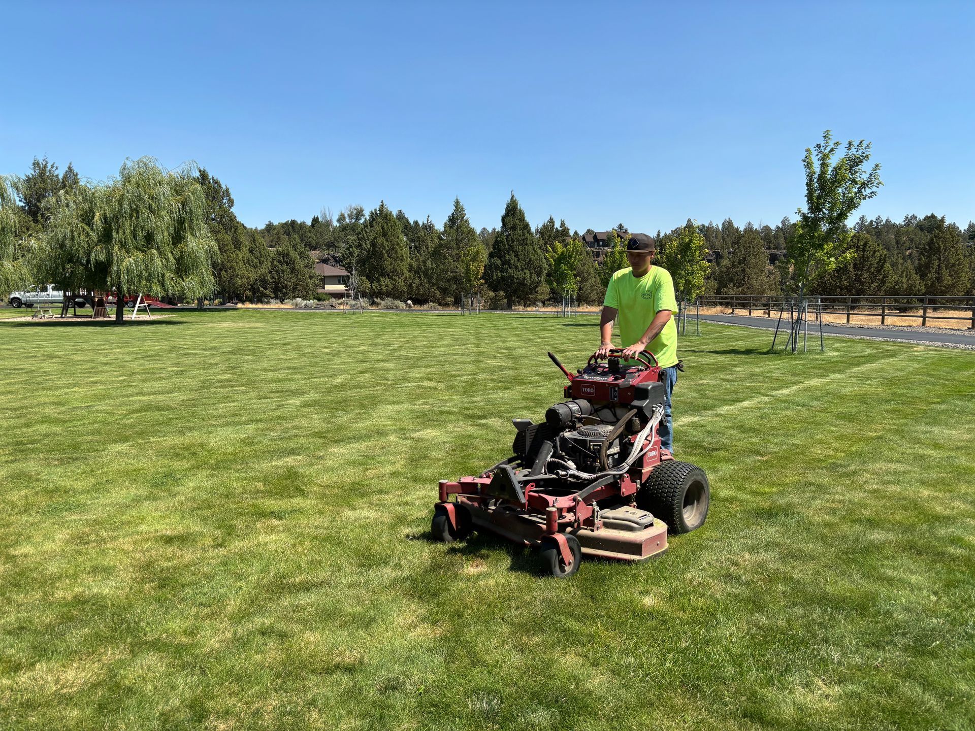 Man mowing a large green lawn with a red riding mower on a sunny day. Trees and a park are in the background.
