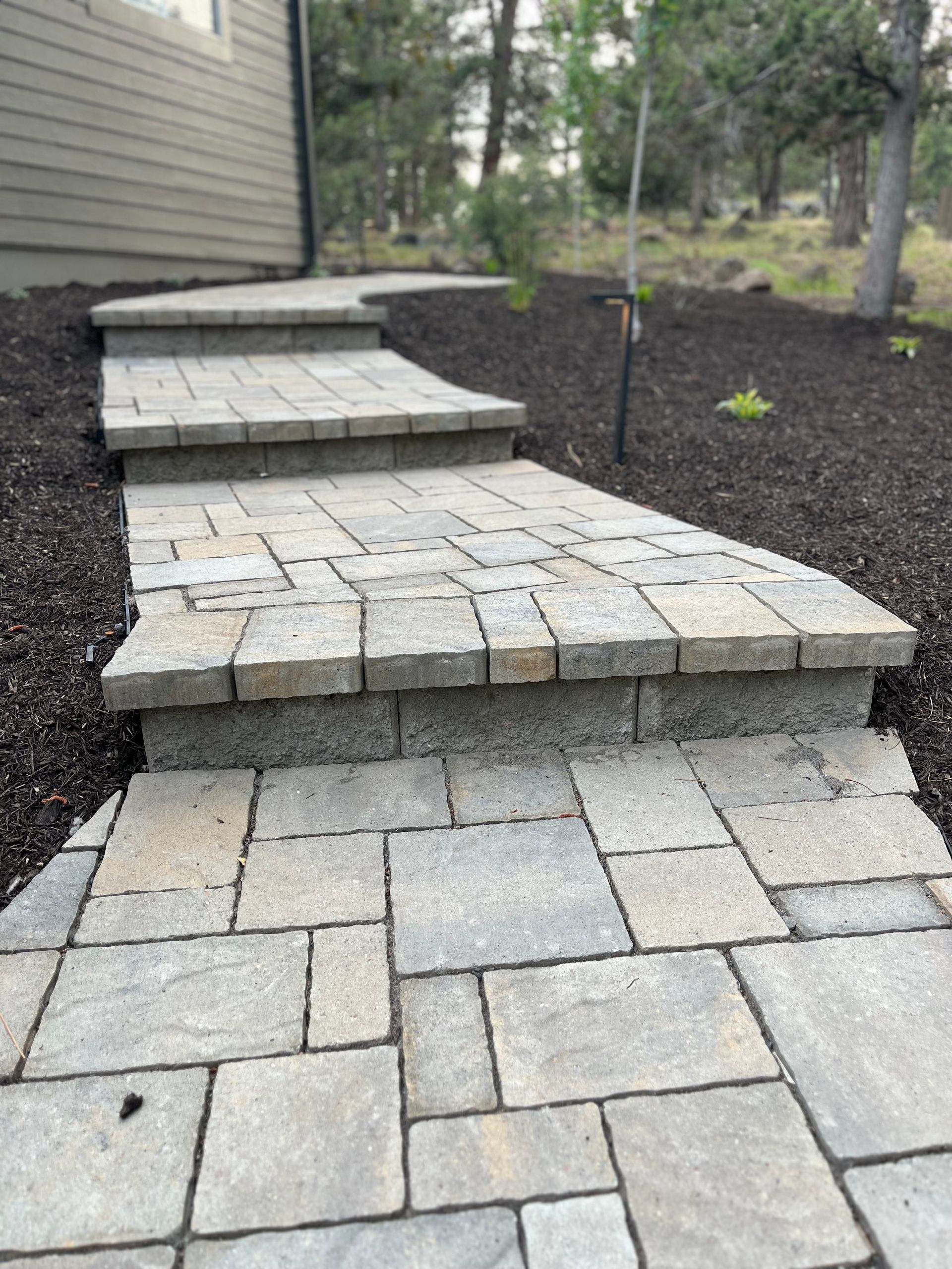Stone steps leading up a pathway, bordered by dark mulch and a light-colored building. Forest in the background.