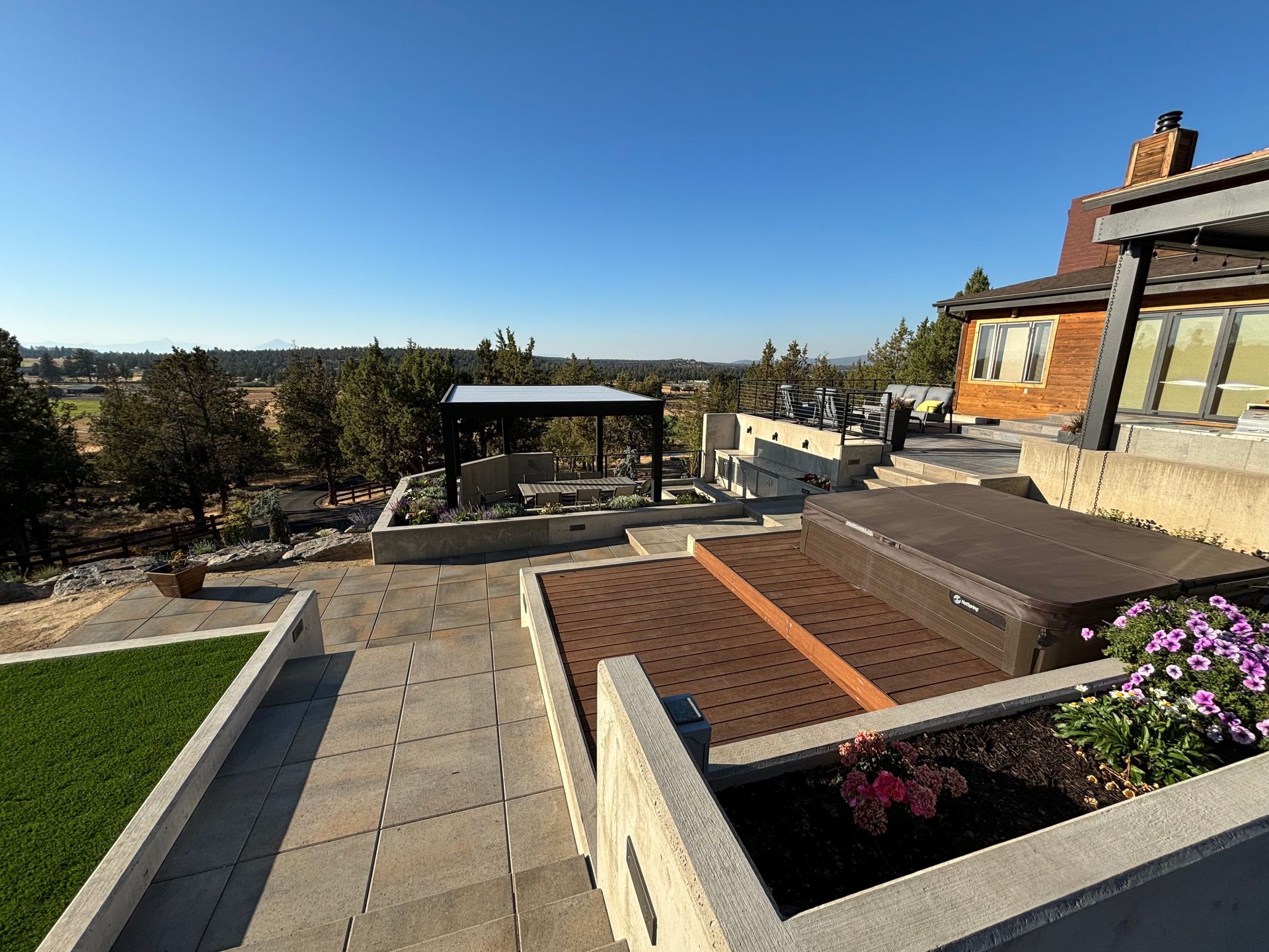 A sunny outdoor patio with a view, featuring raised beds, a pergola, and a house with a brick exterior.
