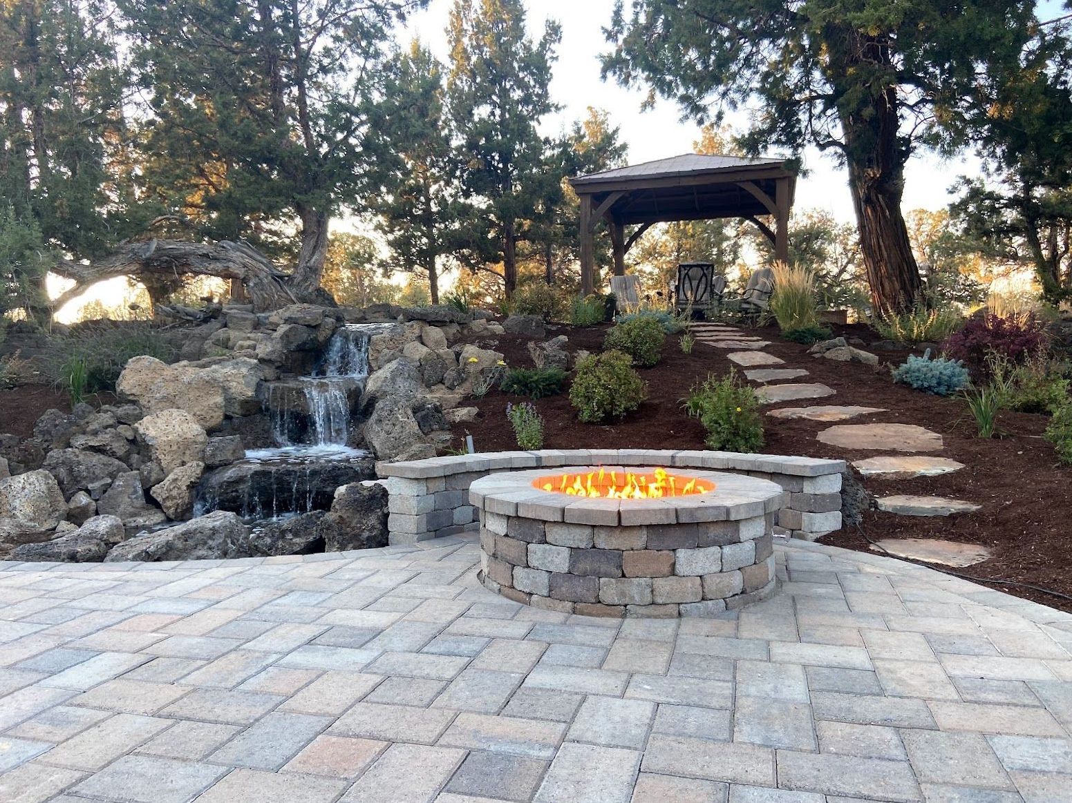 A backyard with a stone fire pit, small waterfall, stepping stones, and a gazebo in the background.