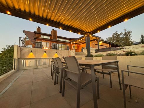 Outdoor patio with dining table and chairs under a pergola. Warm lighting illuminates the seating area and the home in the background.