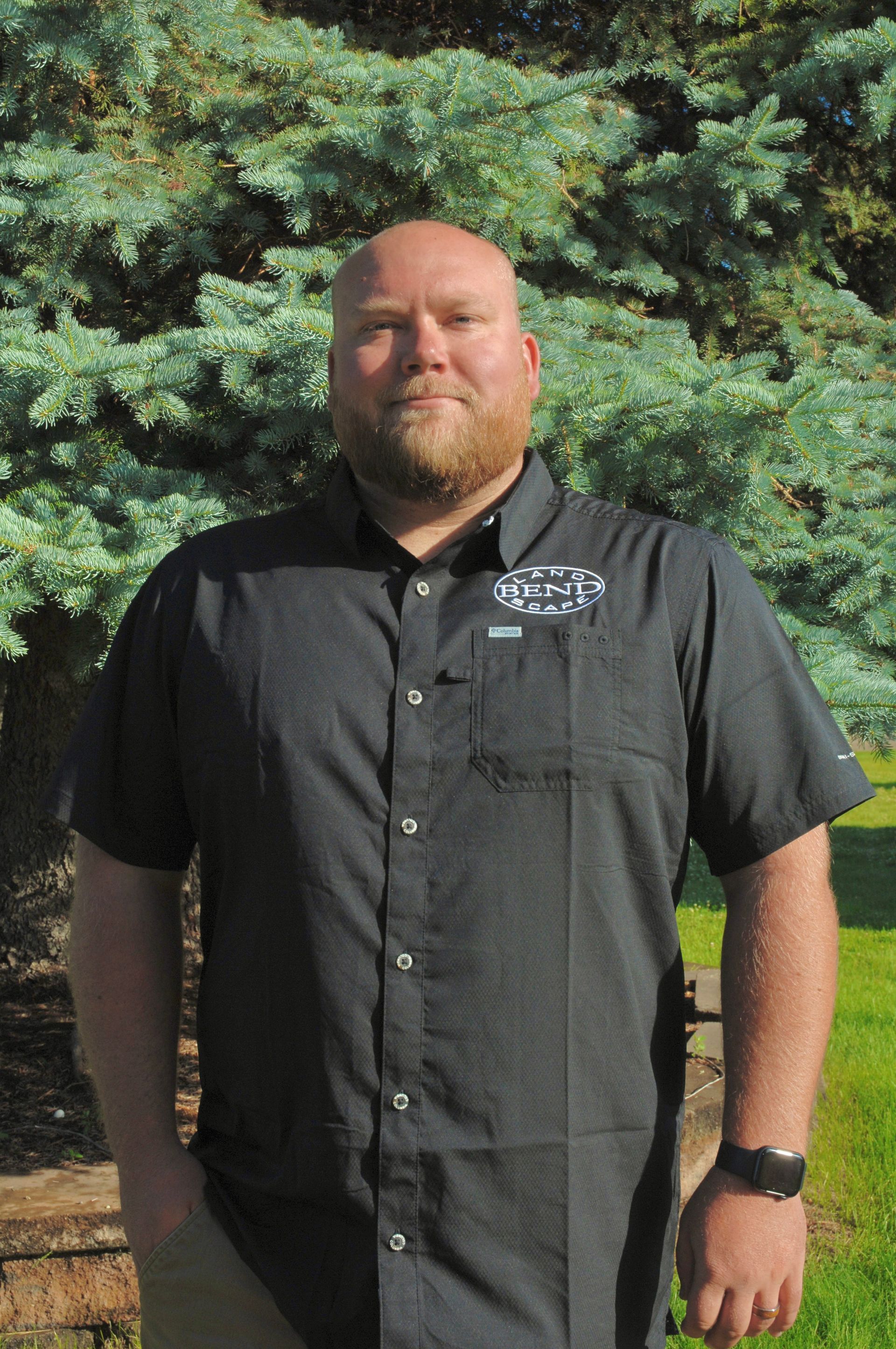 A man with a beard, wearing a black button-up shirt, stands in front of a blue-green bush outdoors.