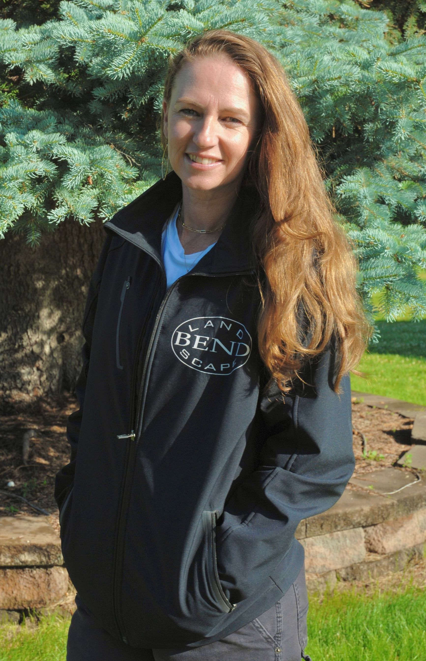 Woman with long curly hair wearing a dark jacket stands in front of a blue-green tree. She is smiling.