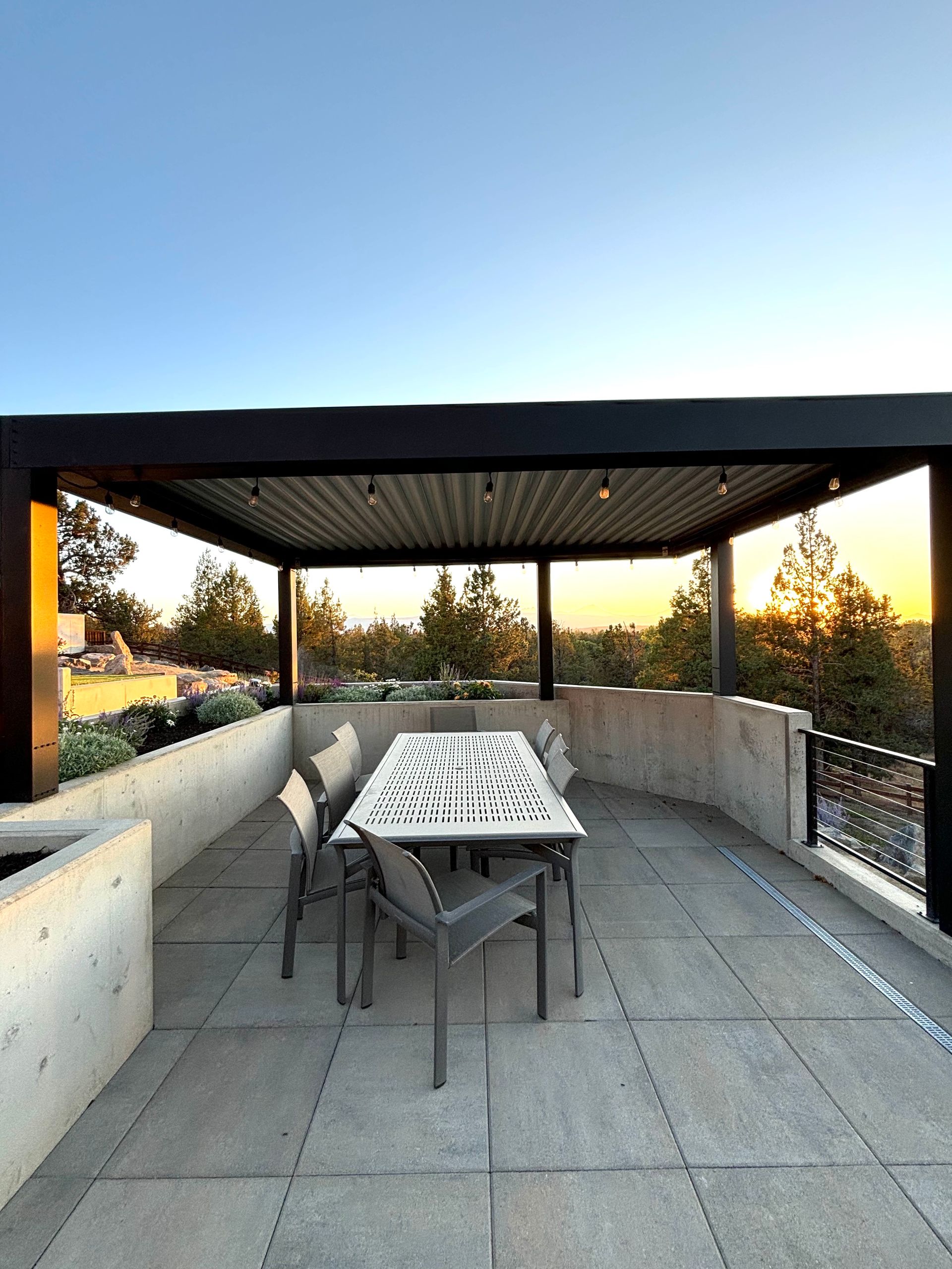 An outdoor dining area with a modern pergola. A table is set with chairs, against a concrete wall overlooking trees at sunset.