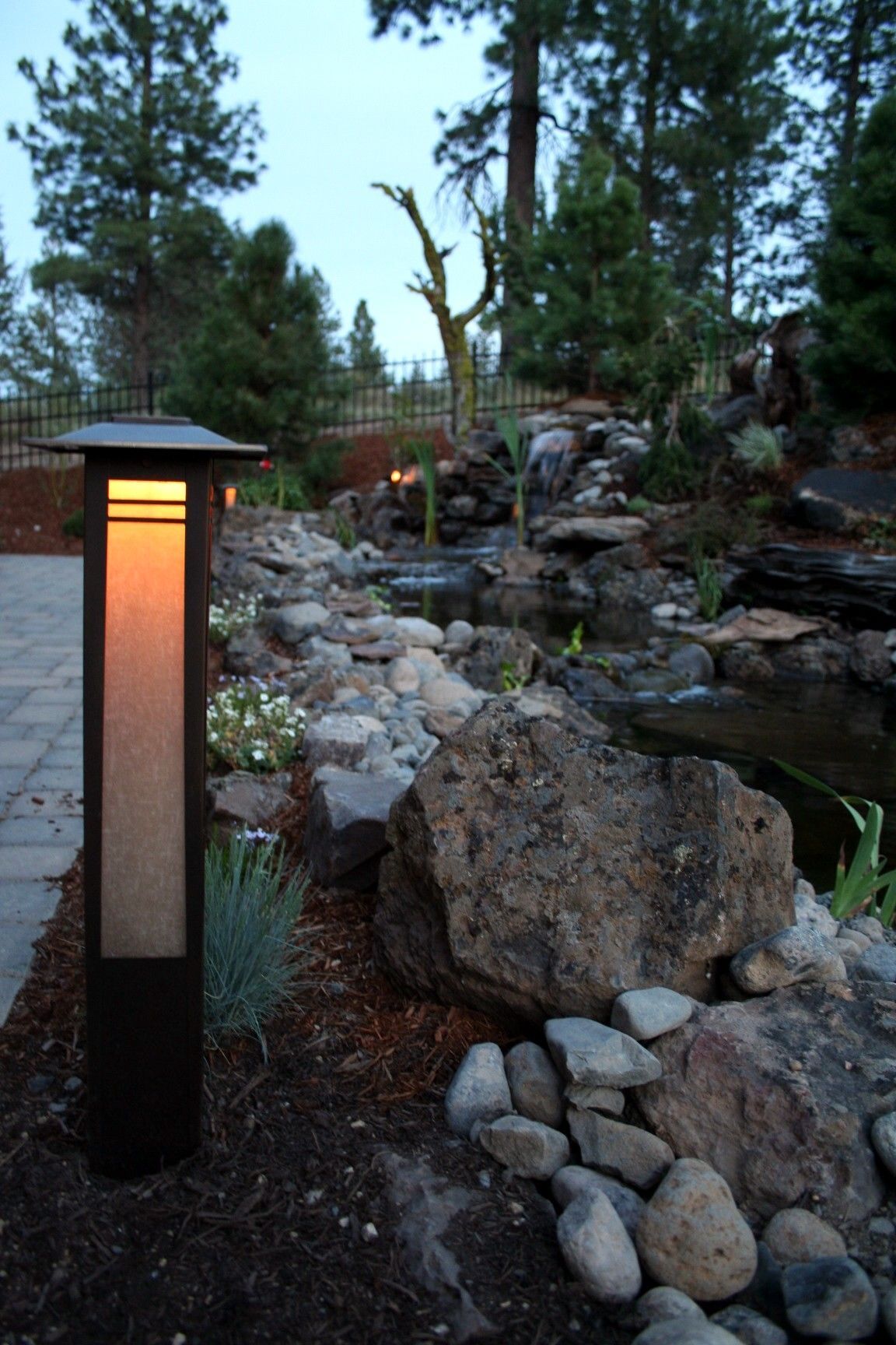 A glowing outdoor lamp illuminates a rocky pond at dusk. Landscaping includes large boulders and greenery.