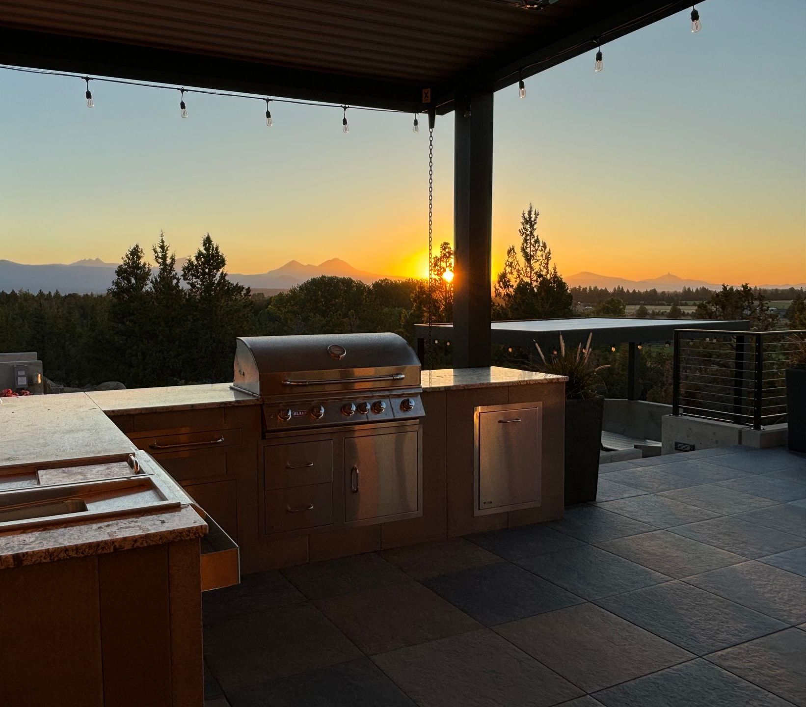 A sunset over mountains viewed from an outdoor kitchen with a built-in grill.