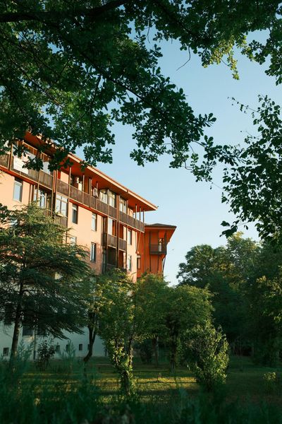 Orange building with wooden balconies, framed by green trees. Blue sky peeks through the foliage.