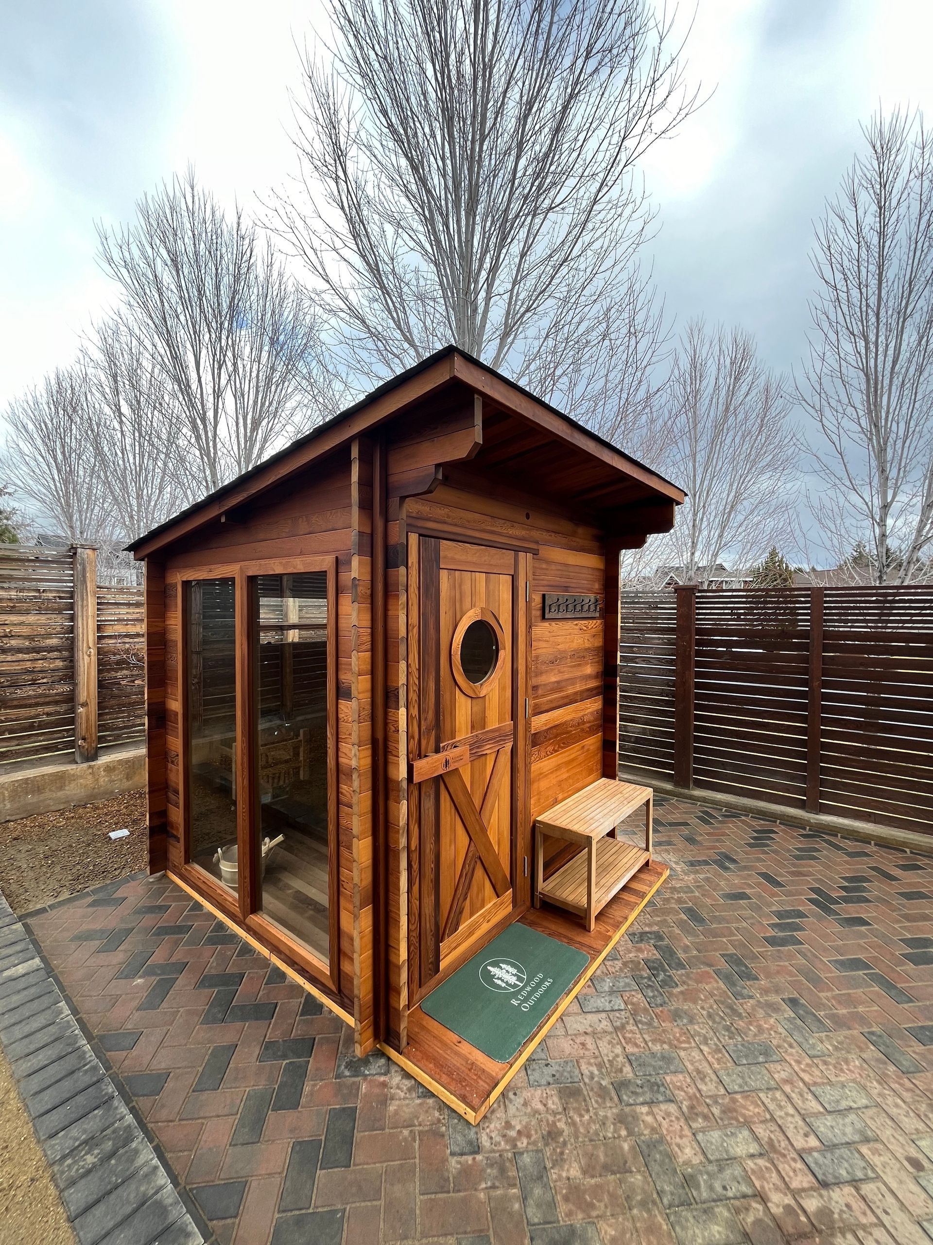 Wooden sauna building on a brick patio with a small bench, in a snowy, outdoor setting.