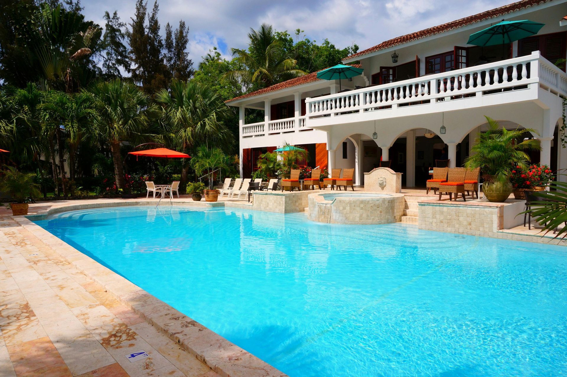 A large, bright blue swimming pool in front of a two-story white house with balconies, surrounded by trees and a sunny sky.