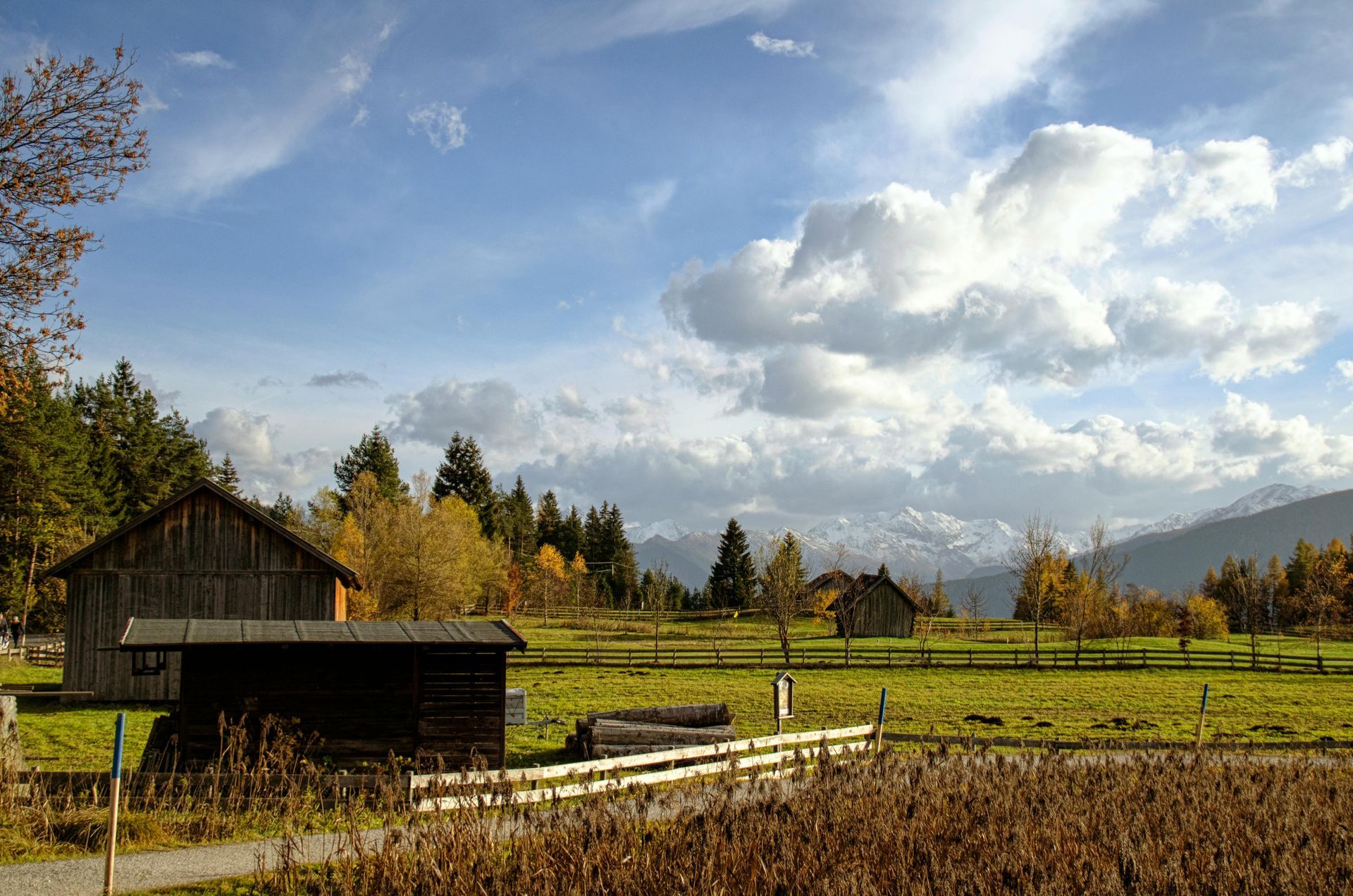 Wooden barns and fields under a blue sky with fluffy white clouds, trees, and distant snow-capped mountains.