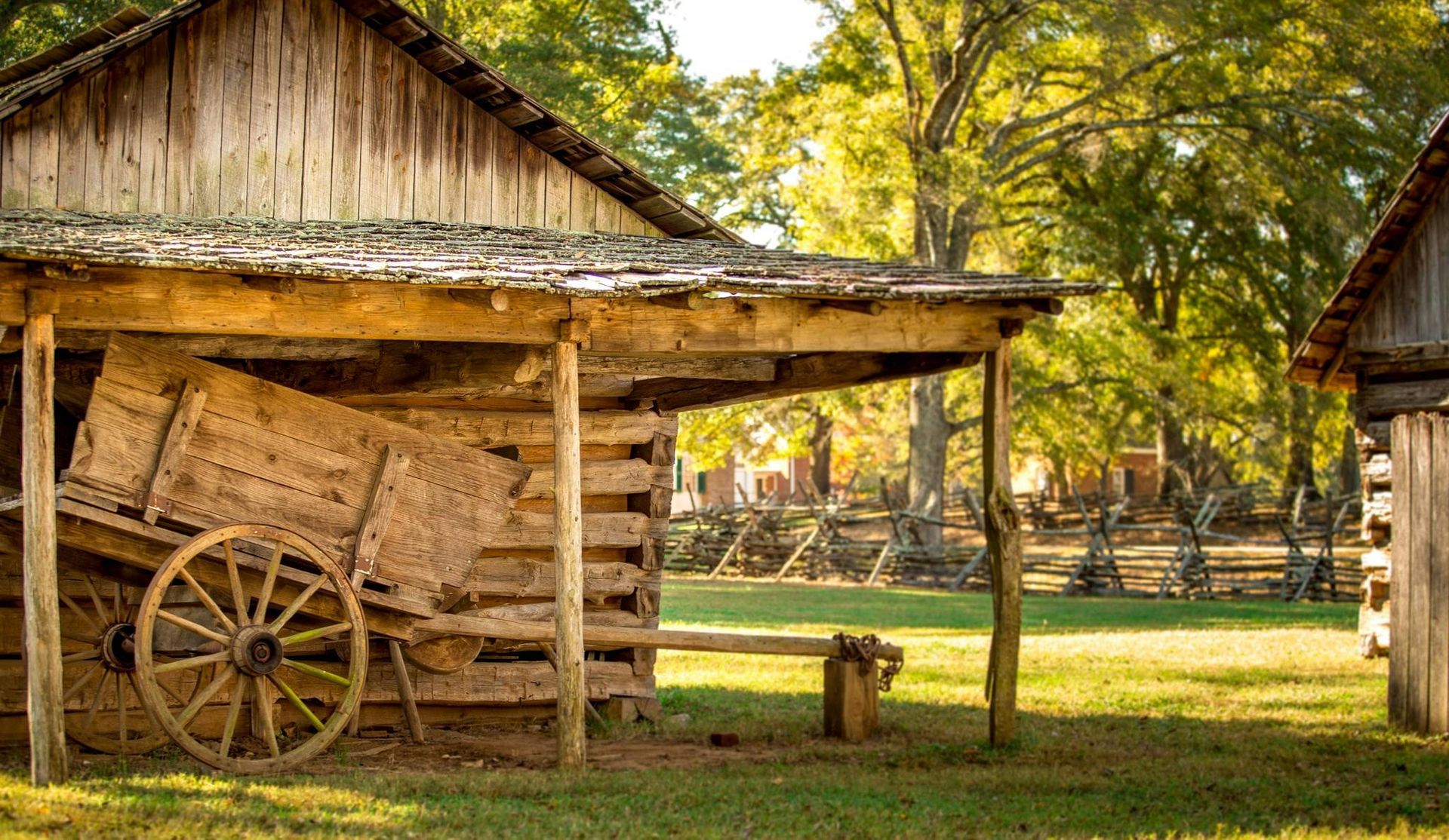A rustic wooden shelter with a weathered cart sits in a grassy field. Sunlight illuminates the scene with trees in the background.