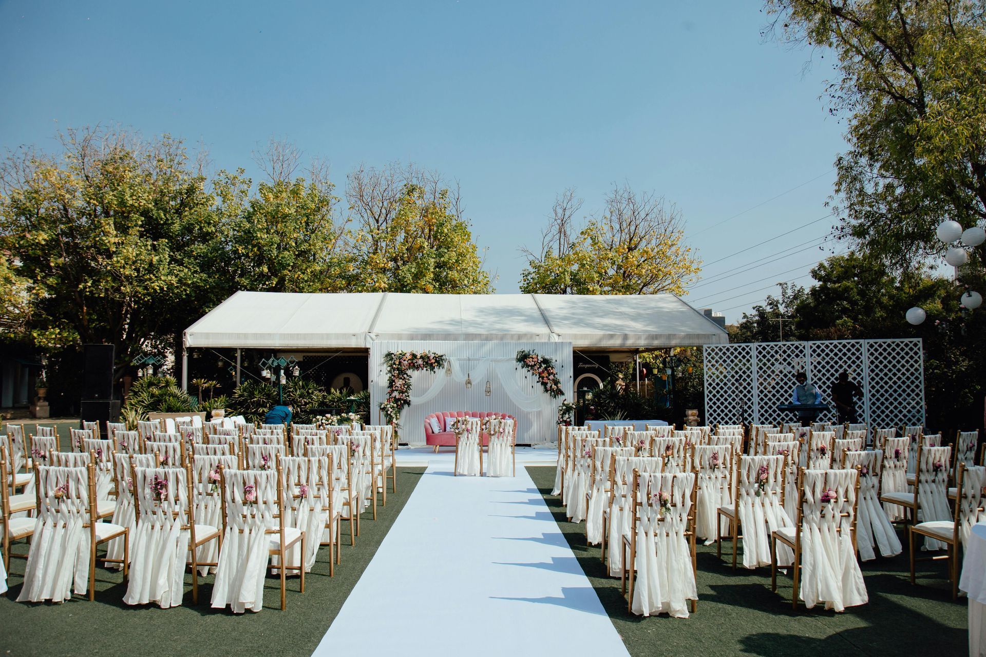 Outdoor wedding ceremony setup with rows of chairs facing an altar under a white tent, on a green lawn under a blue sky.
