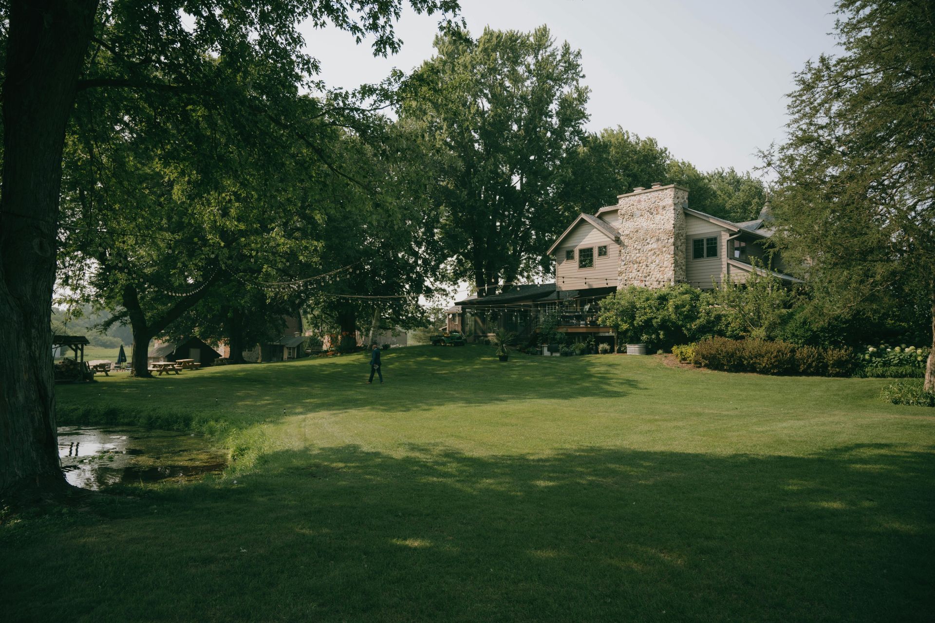 Stone house with turrets set among trees, grassy yard, and a small pond. Sunlight filters through the leaves.