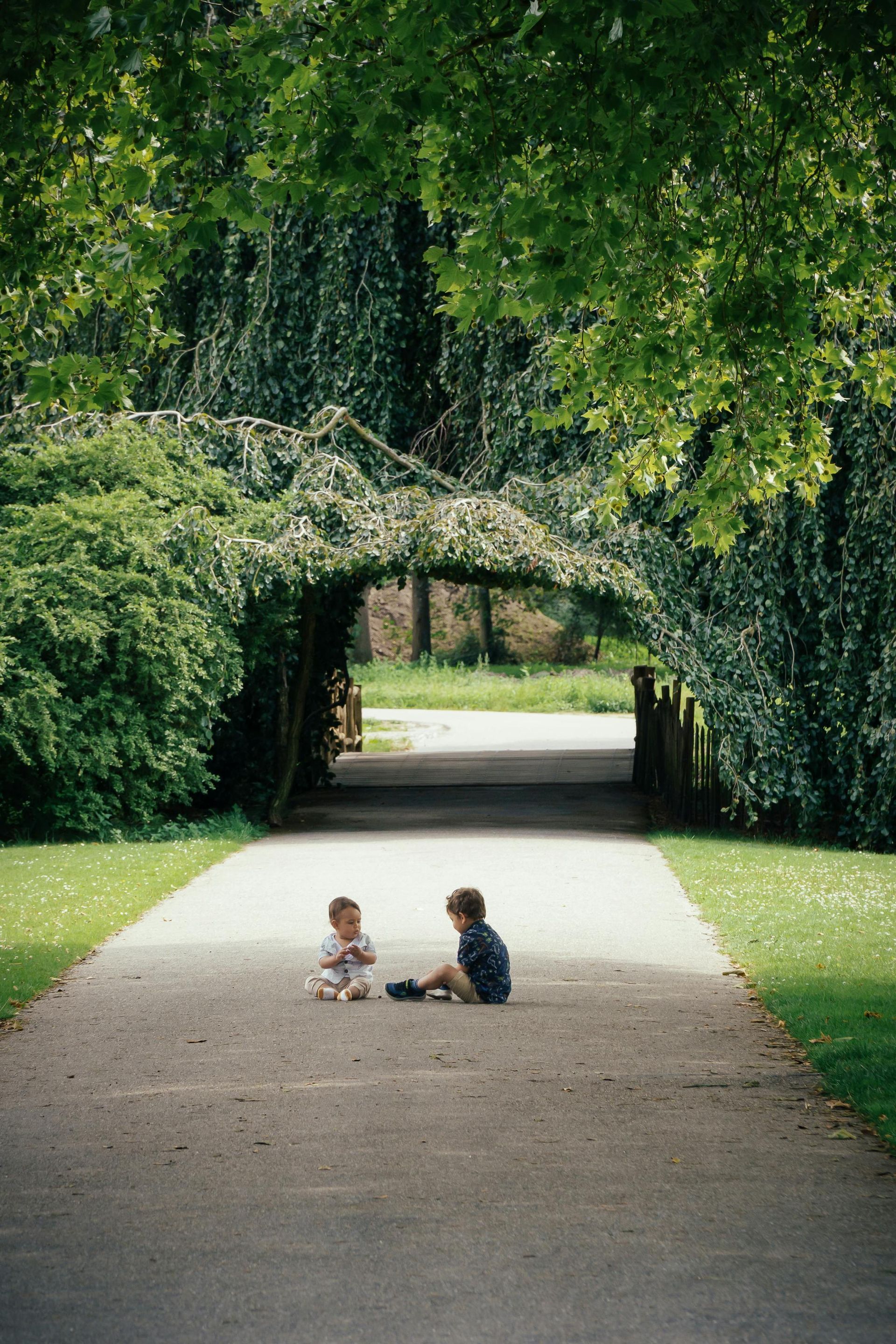 Two children sit on a paved path leading through a leafy archway in a park. Lush green foliage surrounds them.