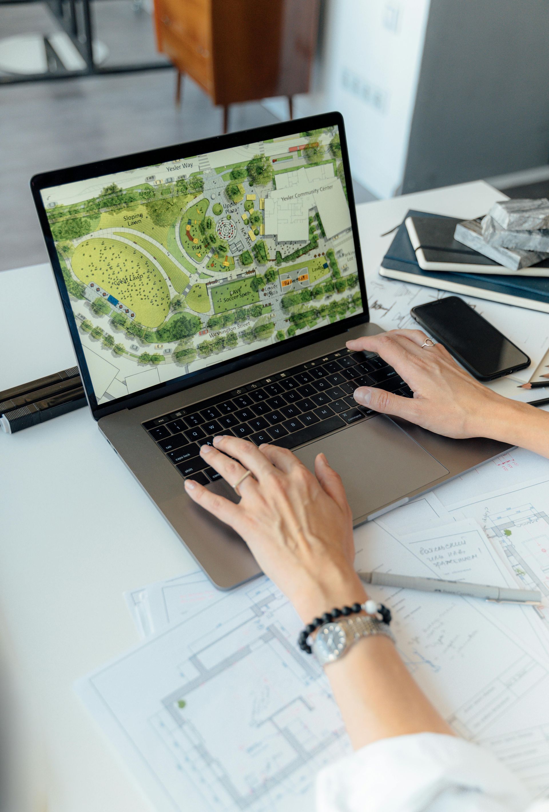 Person working on a laptop displaying a landscape architectural design, surrounded by blueprints and a smartphone on a white desk.