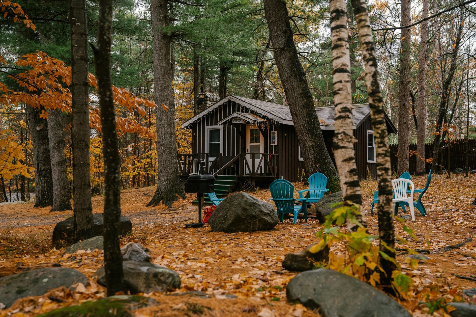 A small cabin nestled among trees with fall foliage, featuring blue and white Adirondack chairs and large rocks.