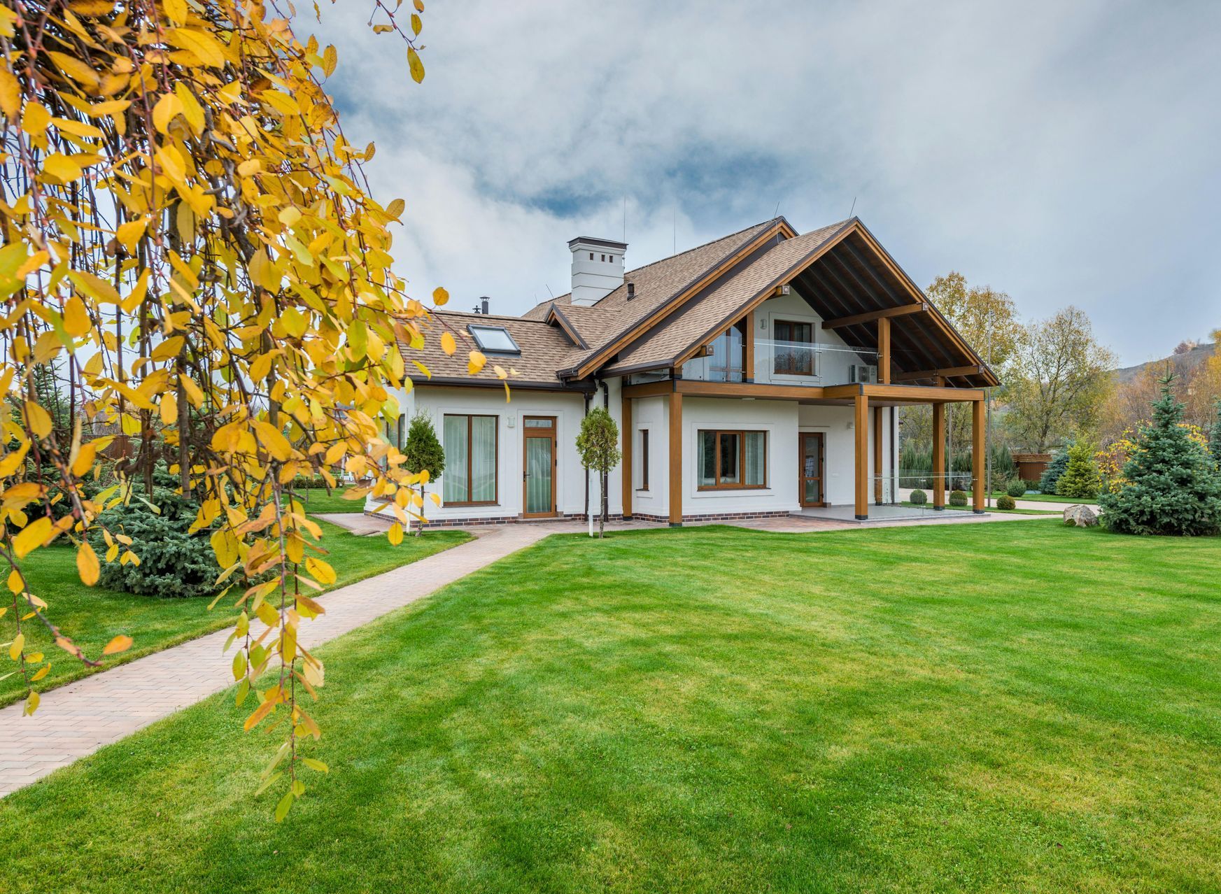 A house with a green lawn and a walkway is surrounded by trees with yellow fall foliage.