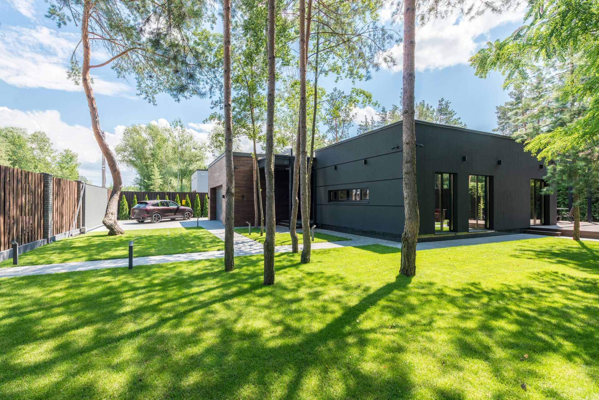 Modern black house with large windows and lush green lawn, trees, and a car in the driveway on a sunny day.