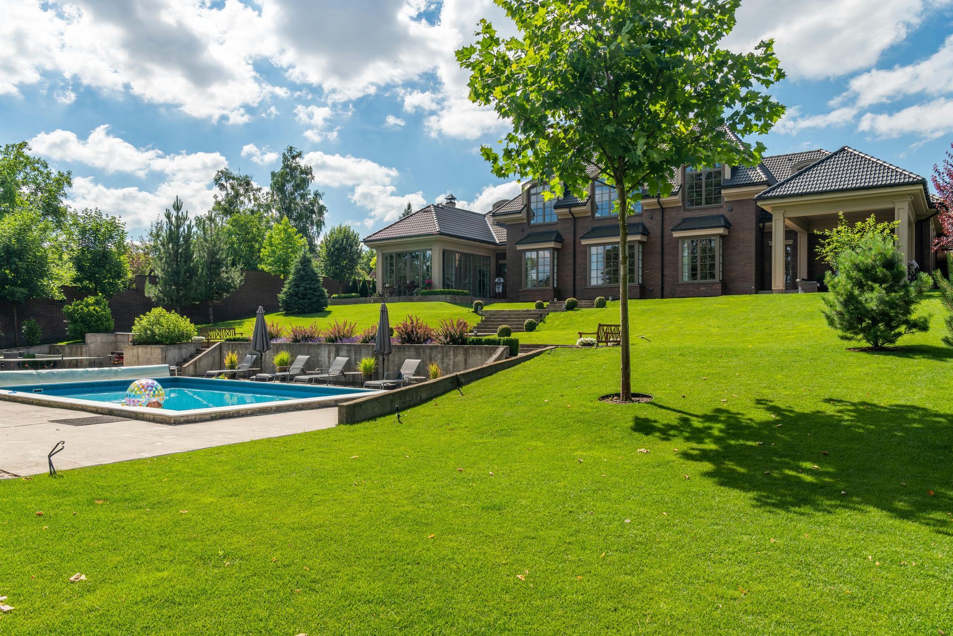 A luxury home with a lush green lawn, swimming pool, and blue sky. A small tree is in the front yard.
