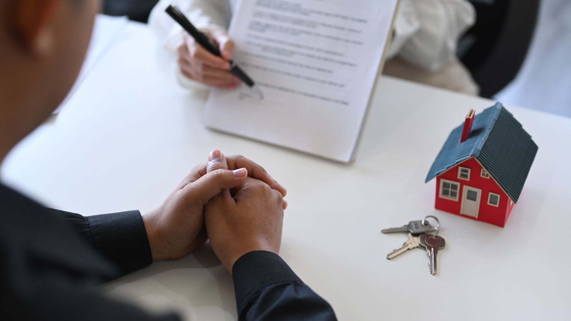 Un hombre está firmando un documento junto a una maqueta de casa y unas llaves.
