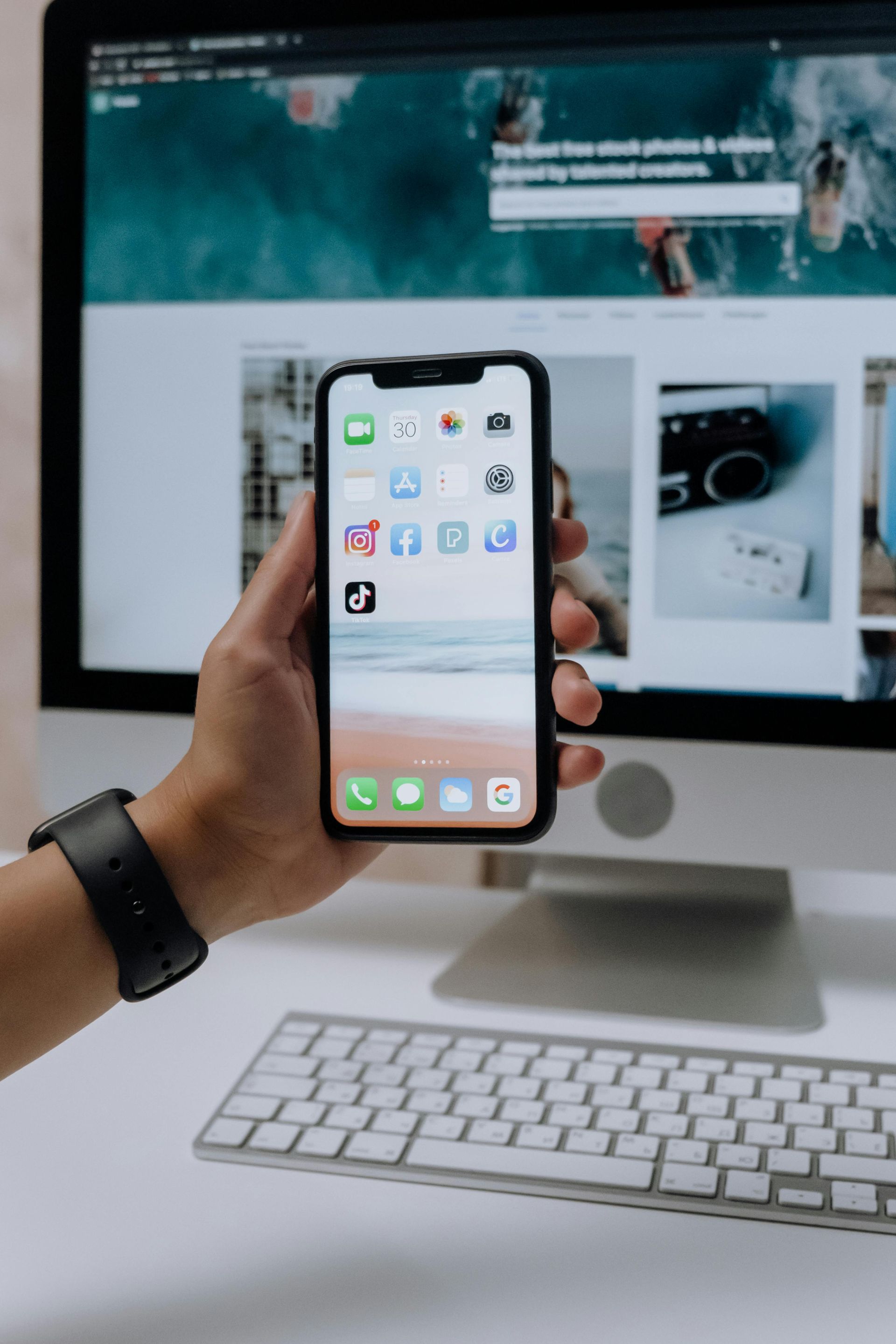 A person is holding a cell phone in front of a computer.