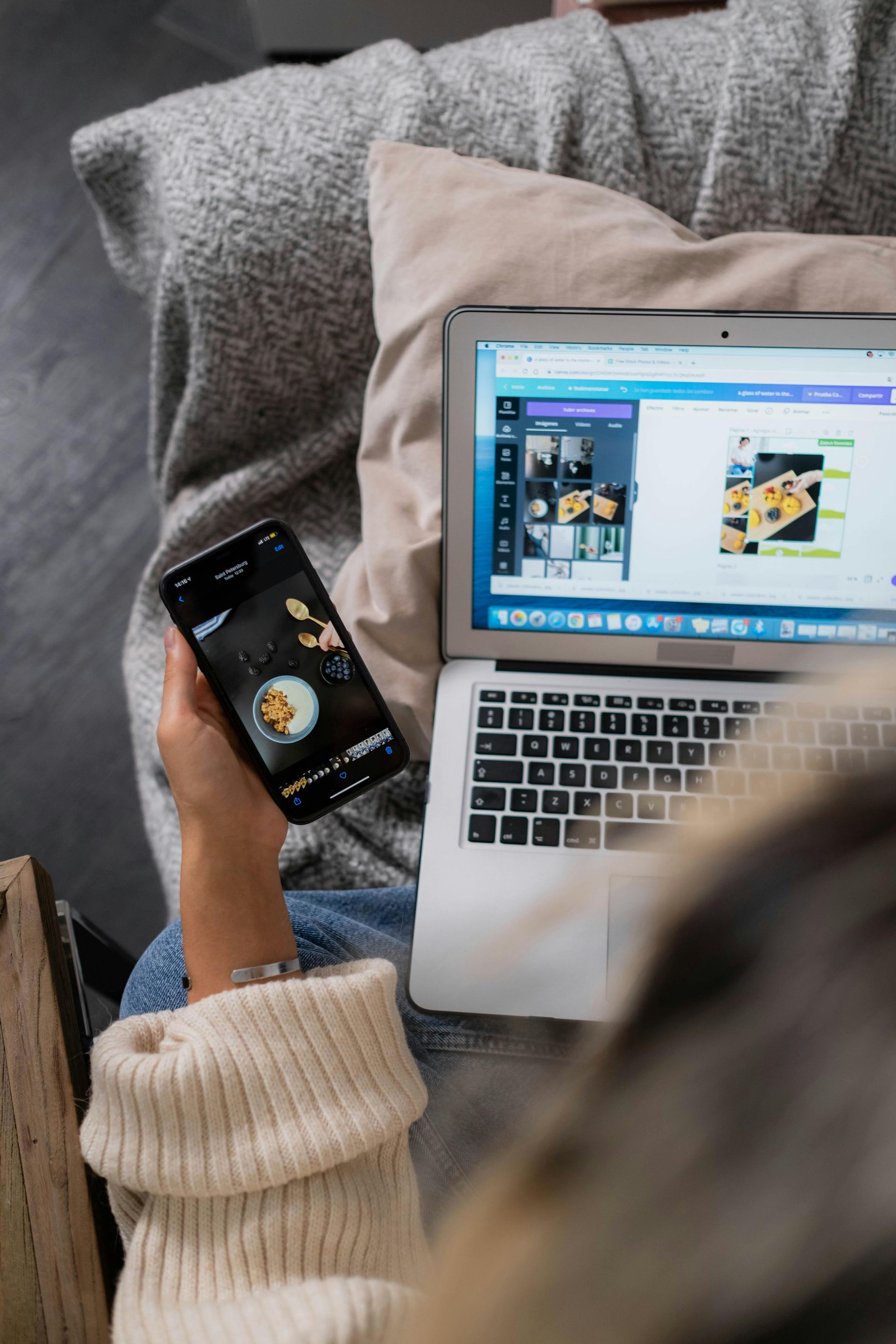 A woman is sitting on a couch holding a cell phone and a laptop doing research in ho to make their website better.