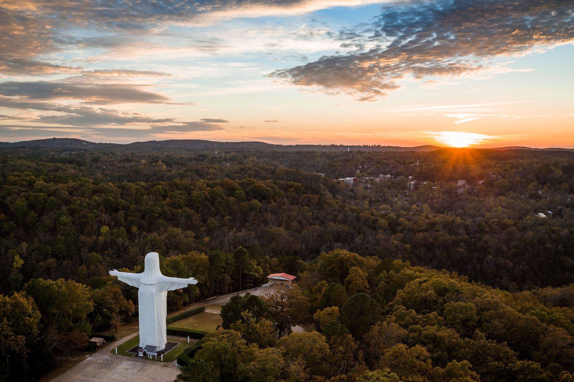 An aerial view of a statue of Jesus surrounded by trees at sunset.