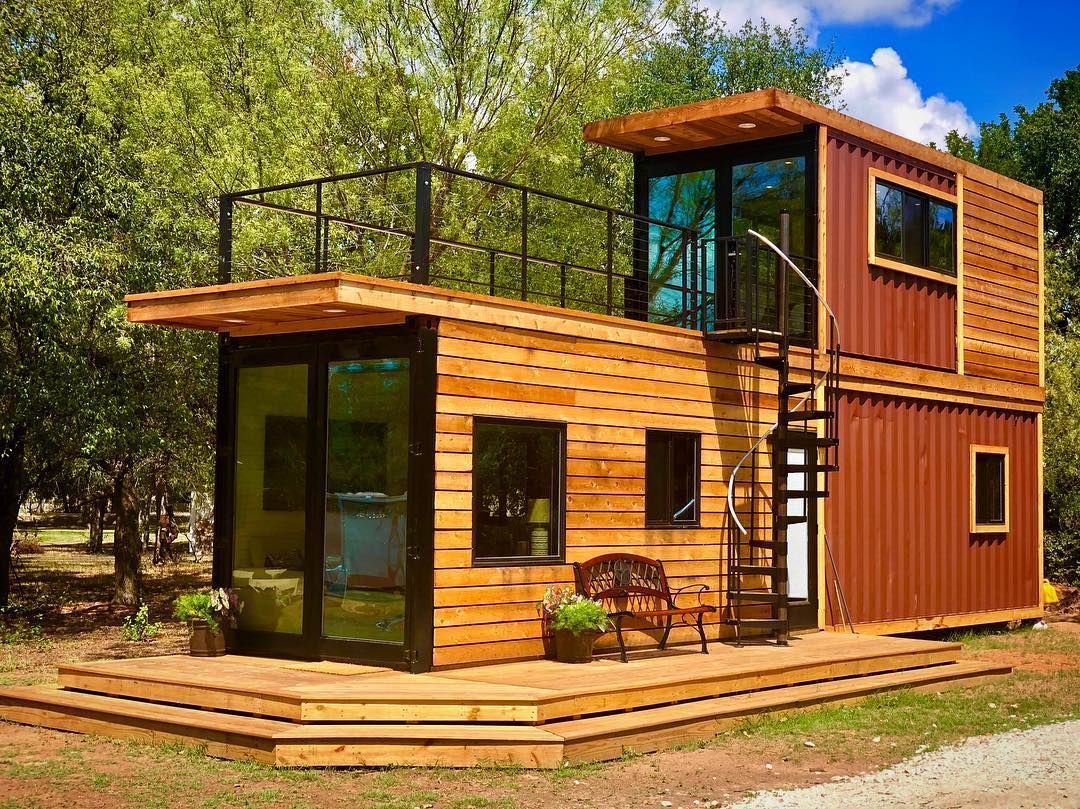 Two-story container home with wooden siding, deck, and metal spiral staircase. Set in a green, outdoor environment.
