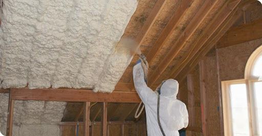 Person spraying insulation in attic; visible beams and windows.